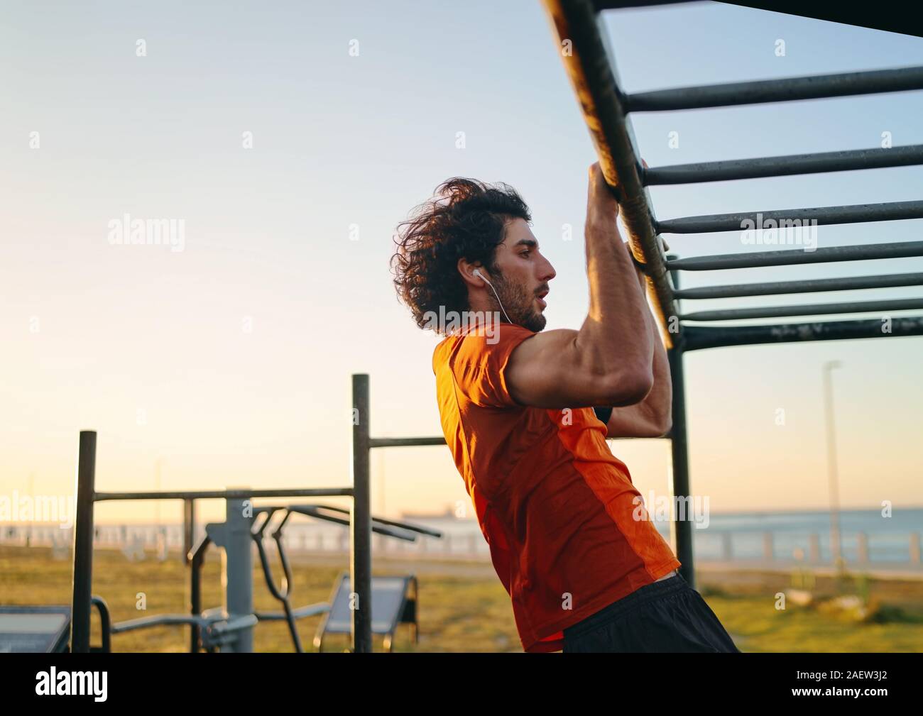 Vue latérale d'un jeune homme fit musculaire avec vos écouteurs dans ses oreilles faisant tirer-se lève à la salle de sport en plein air dans le parc Banque D'Images