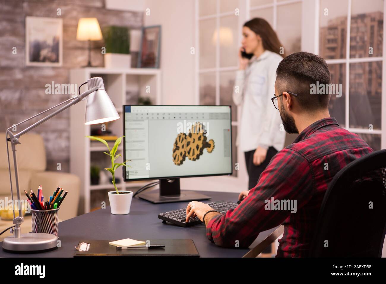 Vue arrière de l'ingénieur mâle dimensions un prototype à l'aide du logiciel de CAO mécanique dans la salle de séjour. Petite amie à l'arrière-plan. Banque D'Images
