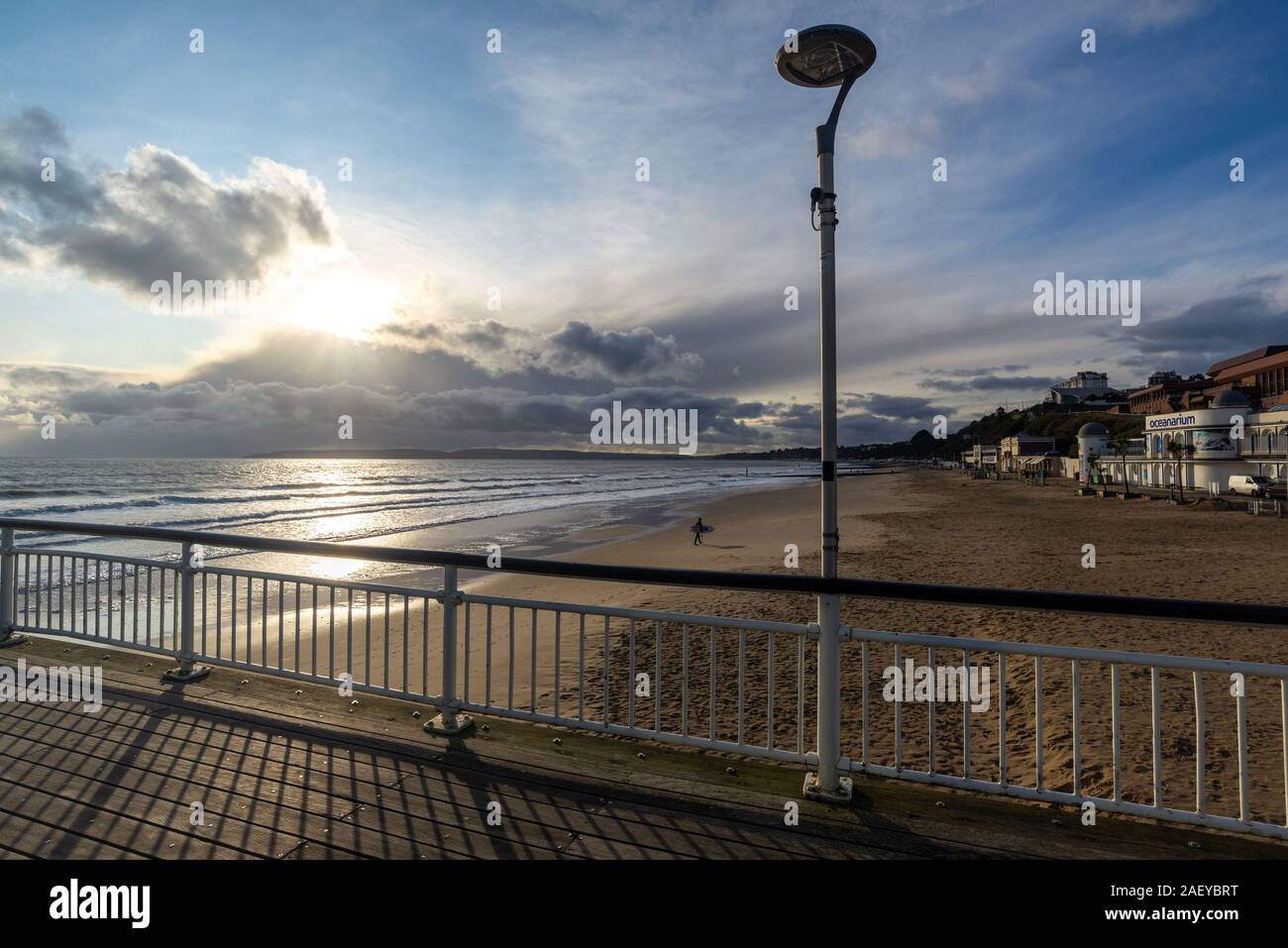 En hiver, de Bournemouth Dorset, UK, décembre 2019. Froid avec des averses de pluie, ensoleillée, avec un vent fort sur la côte sud de l'après-midi. Soleil et nuages orageux sur l'île de Purbeck. Un hardy surfer carrying un surf sur la plage après le surf dans l'eau de mer froide. Banque D'Images