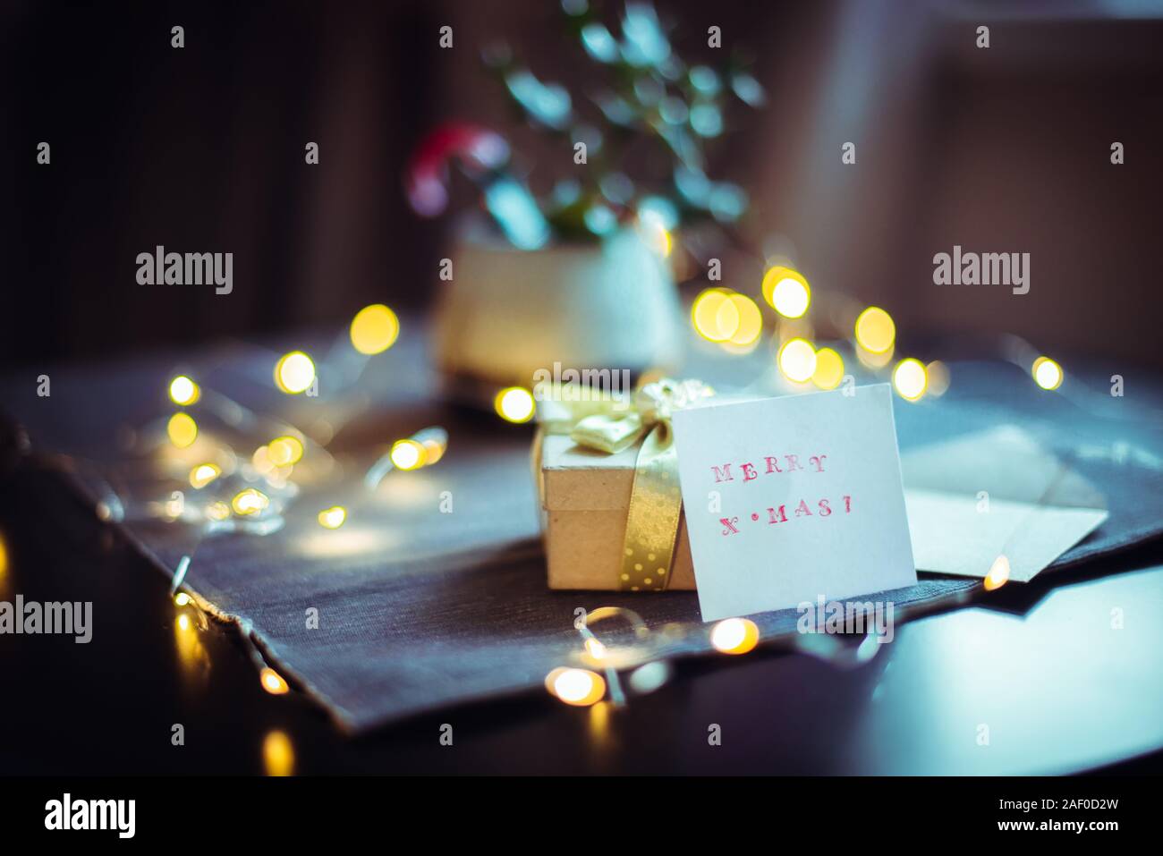 Close up hand made stamp Joyeux Noël, carte vintage boîte-cadeau avec ruban doré et festive avec des lumières composition garland sur serviette gris. Sc sombre Banque D'Images