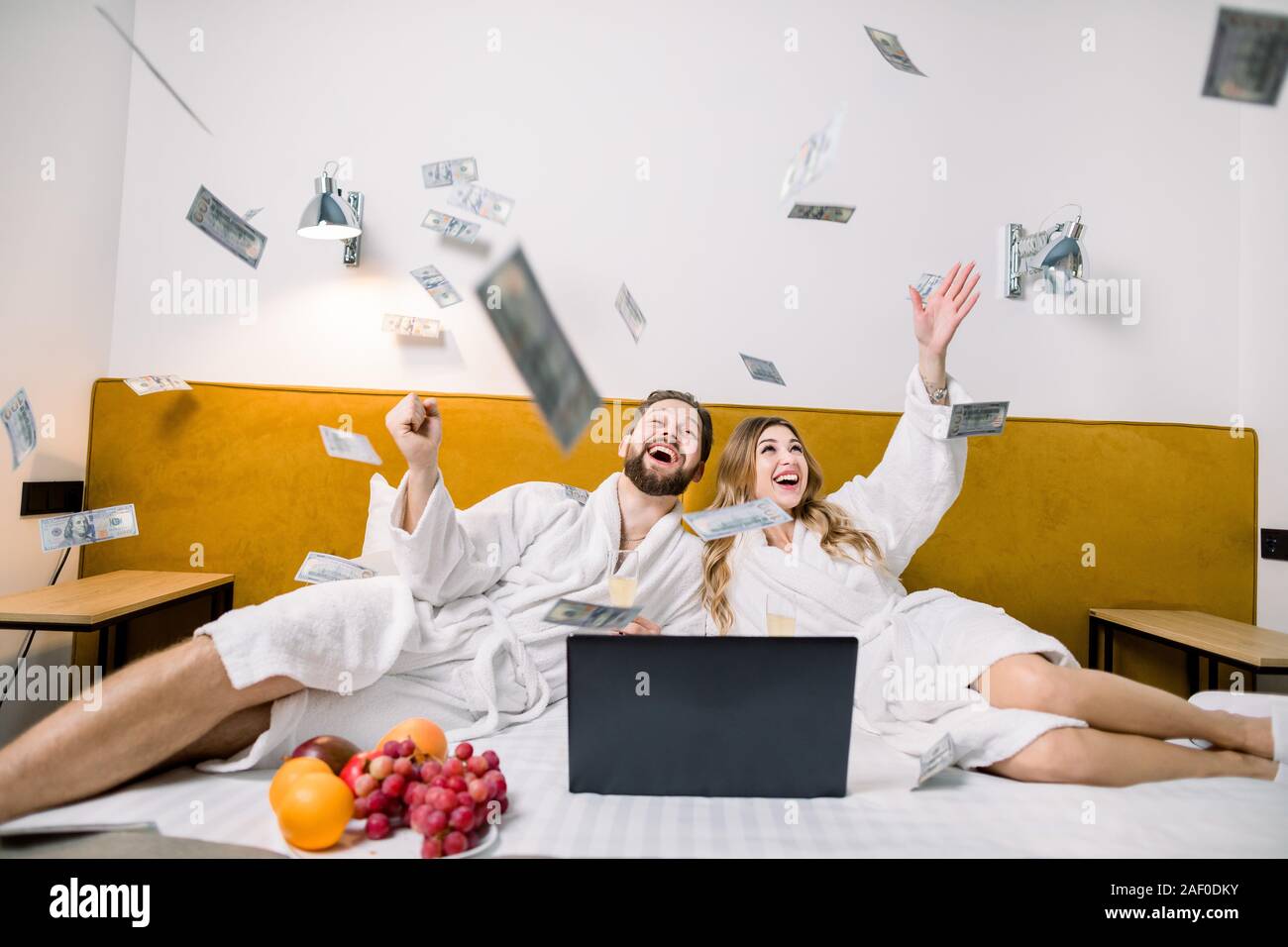 Image d'un jeune beau couple de jeunes mariés jeter de l'argent, tout en étant assis sur le lit dans la chambre d'hôtel, à l'aide d'ordinateur portable et manger des fruits. Banque D'Images