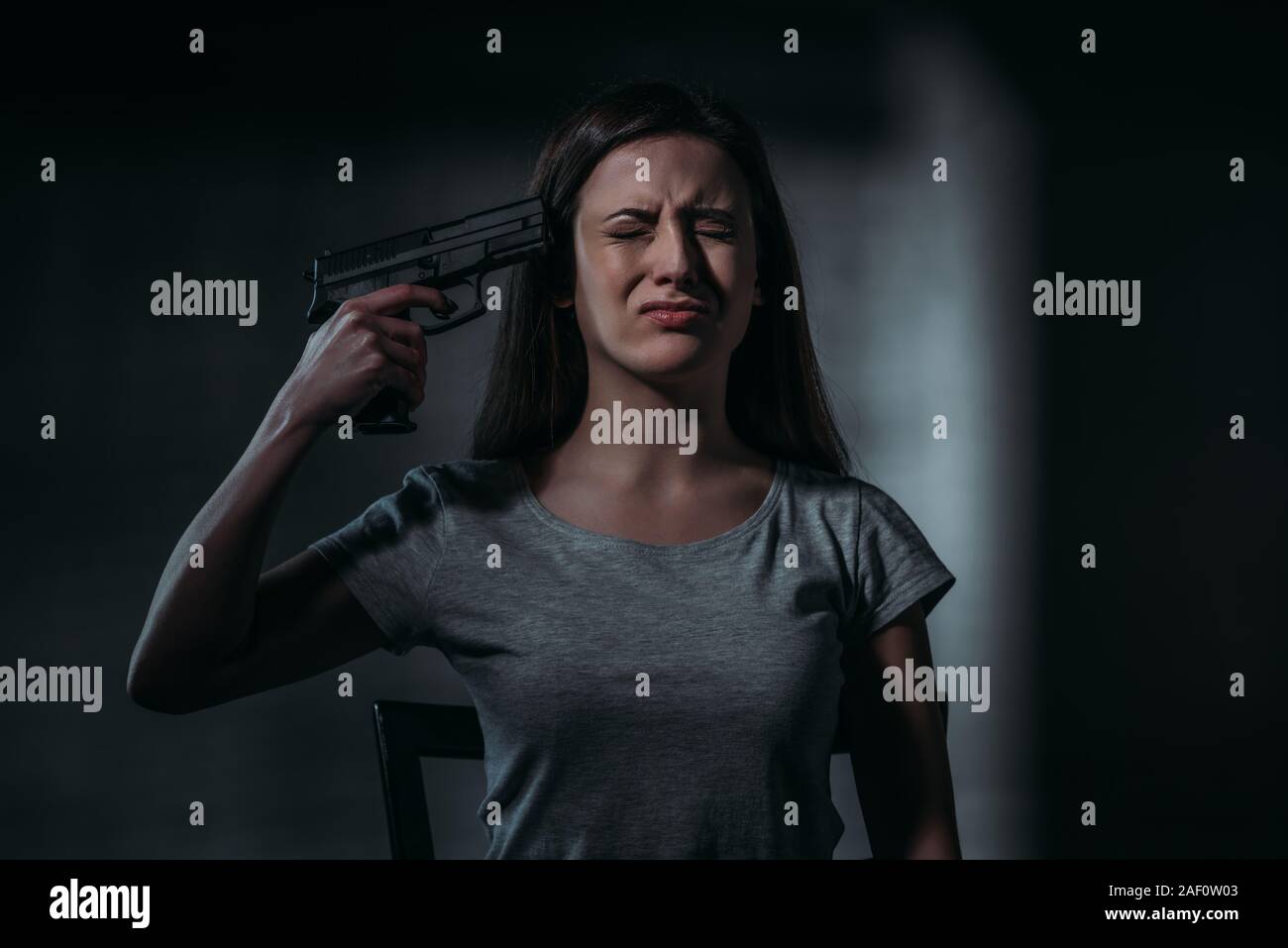 Pleurer, désespéré woman holding gun près de menton sur fond sombre Banque D'Images