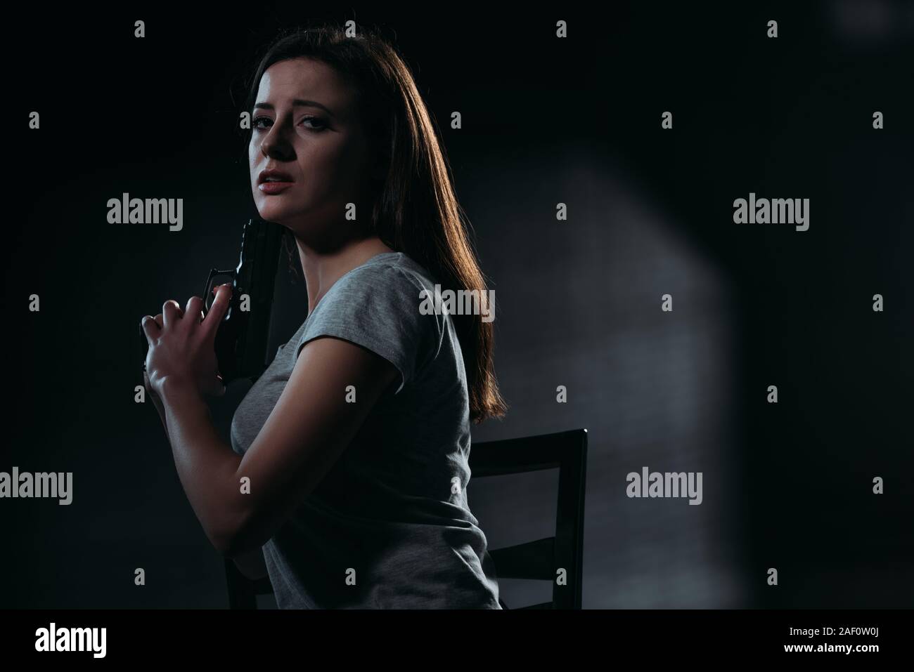 Déprimé woman looking at camera while holding gun près de menton sur fond sombre Banque D'Images