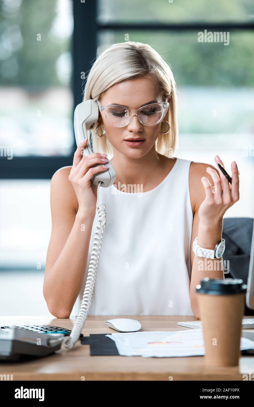 Portrait of businesswoman in glasses looking at documents et parler sur retro phone près de tasse de papier Banque D'Images