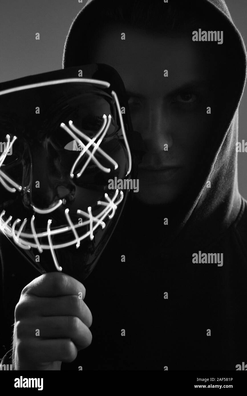 Portrait d'un homme anonyme dans un hoodie noir cachant son visage derrière un masque de néon effrayant. Studio shot. Banque D'Images