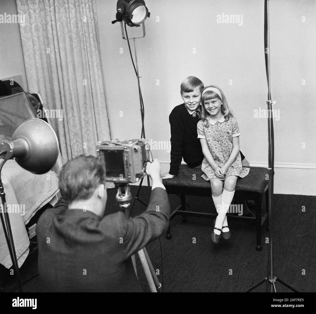 Années 1960, historique, assis à l'intérieur d'un studio photographique, deux enfants, un frère et une sœur, se faisant prendre en photo par un photographe professionnel à l'aide d'un appareil photo sur un trépied, Angleterre, Royaume-Uni. Banque D'Images