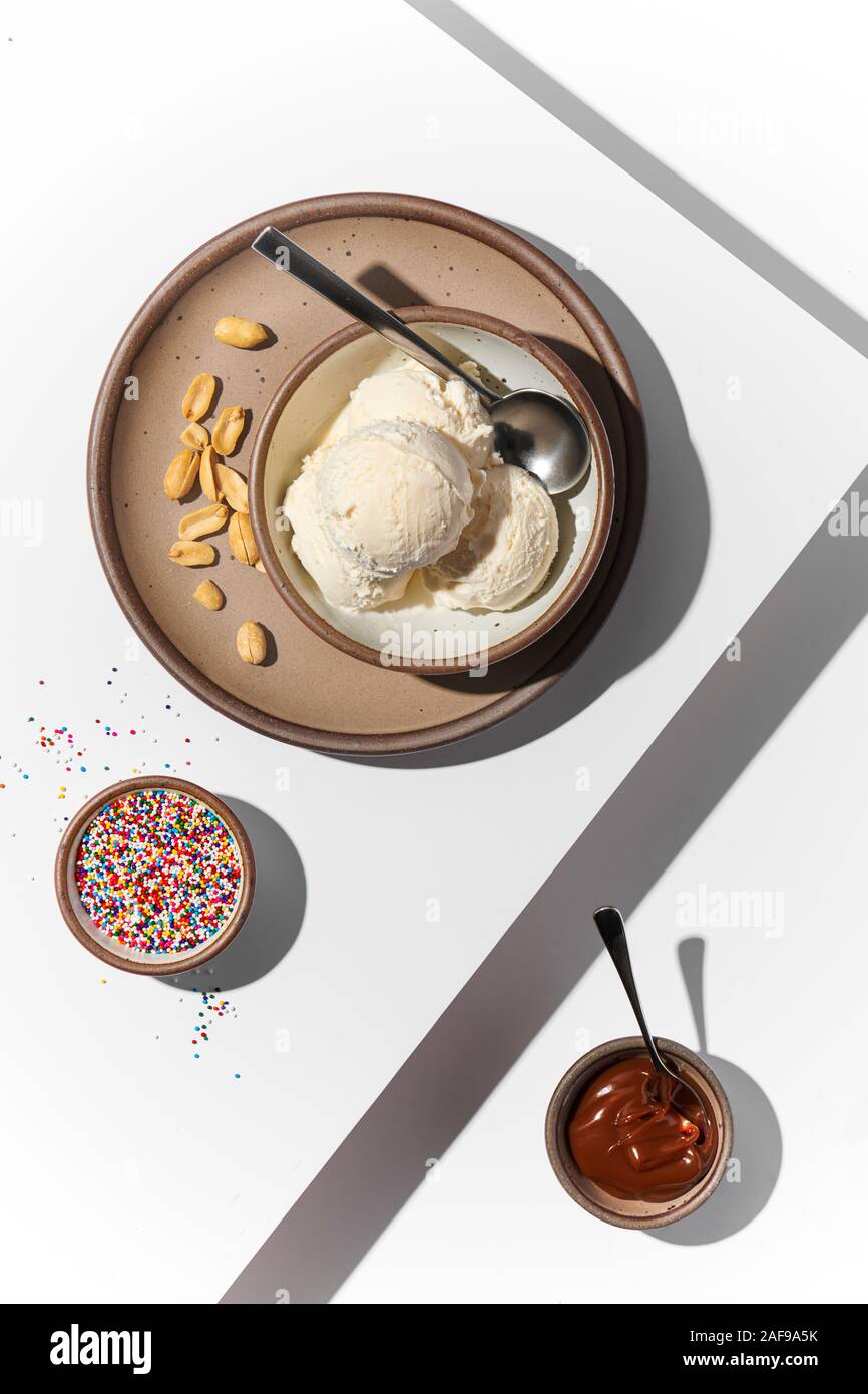 Bol de crème glacée à la vanille avec des petits bols de garnitures, y compris les arachides, rainbow et saupoudre de sucre. Surface blanche avec disque contraste des ombres Banque D'Images