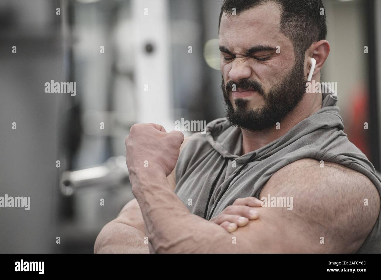 Douleur musculaire blessure concept de jeune homme avec barbe portant un casque sans fil et hoodie holding dommages hid biceps lors d'un entraînement dans la salle de sport Banque D'Images