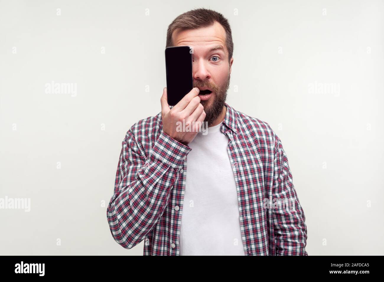 Portrait d'homme barbu surpris en chemise à carreaux occasionnel pour une moitié face avec téléphone mobile et à la recherche en surprise à l'appareil photo, de la technologie et de l'identit Banque D'Images