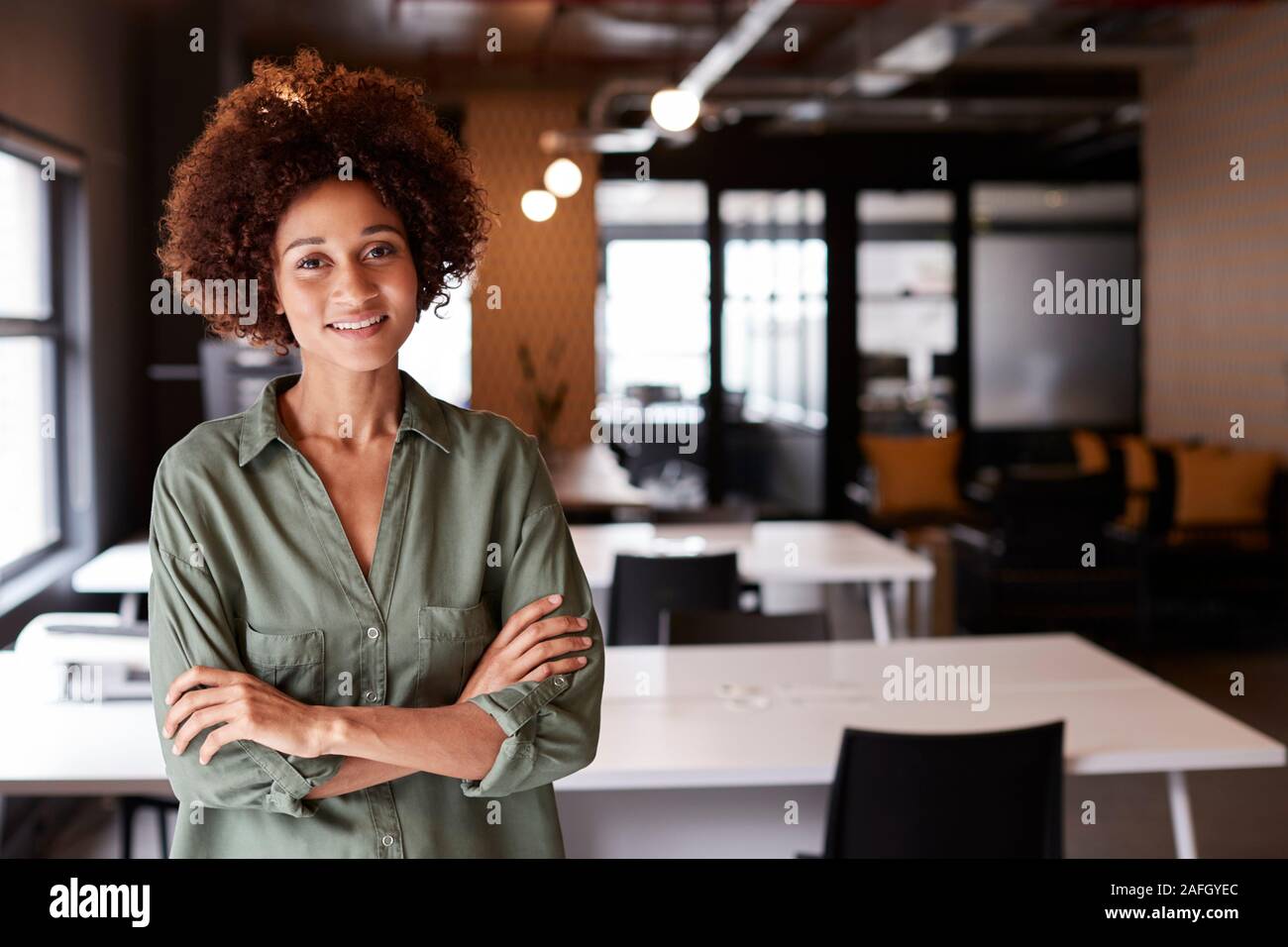 Femelle noire millénaire creative debout dans un bureau en espace ouvert avec les bras croisés smiling to camera Banque D'Images