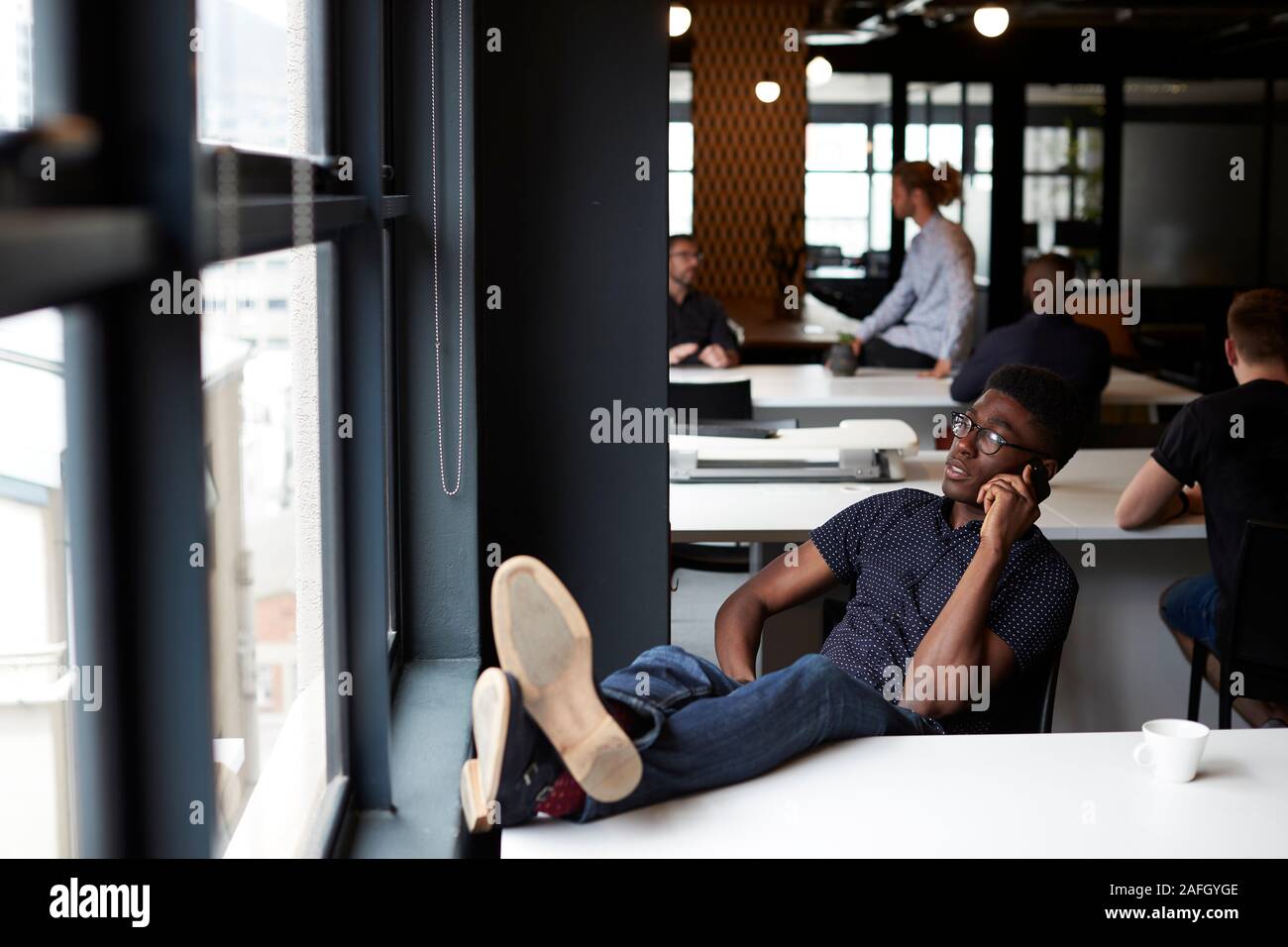 Jeune homme noir creative assis dans un bureau avec ses pieds sur le bureau en utilisant le téléphone, Close up Banque D'Images