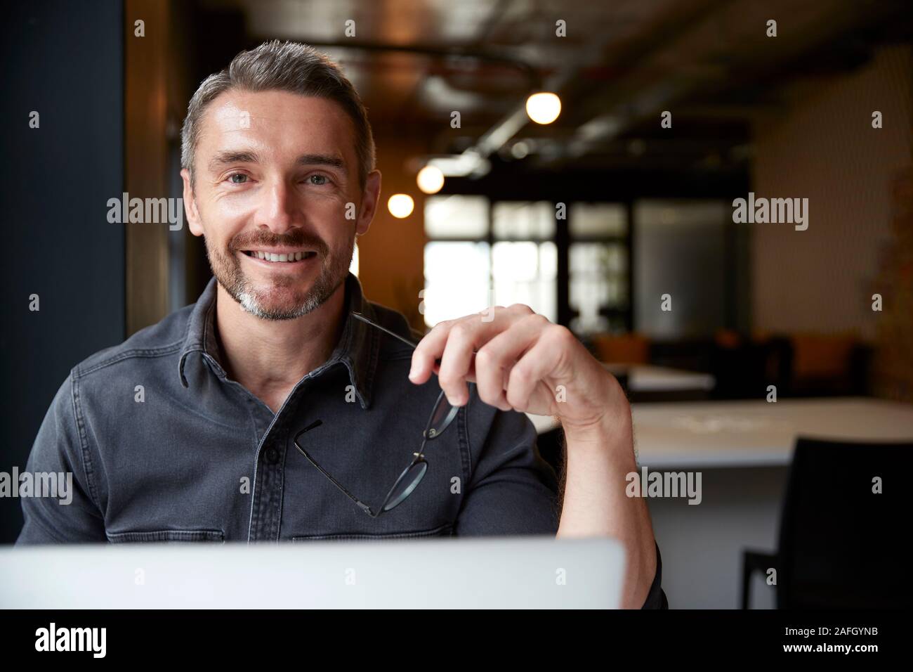 Homme de race blanche d'âge moyen creative sitting at desk in office holding glasses, souriant à la caméra, Close up Banque D'Images