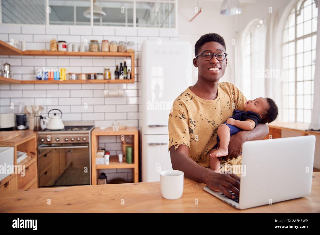 Portrait du père multitâches Holding Baby son sommeil et working on laptop computer in Kitchen Banque D'Images