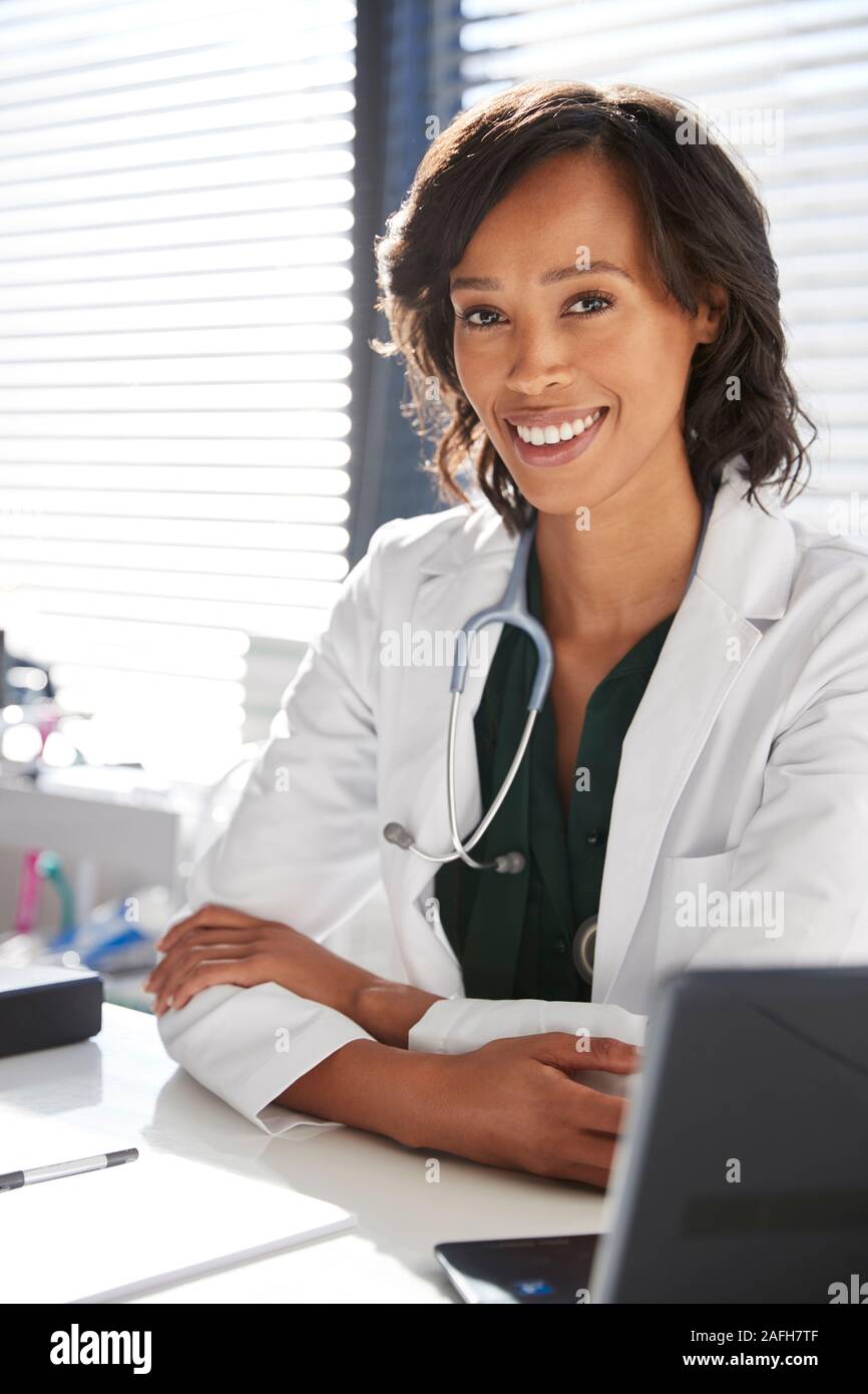 Portrait Of Smiling Woman Wearing White Coat with Stethoscope Sitting Behind Desk In Office Banque D'Images