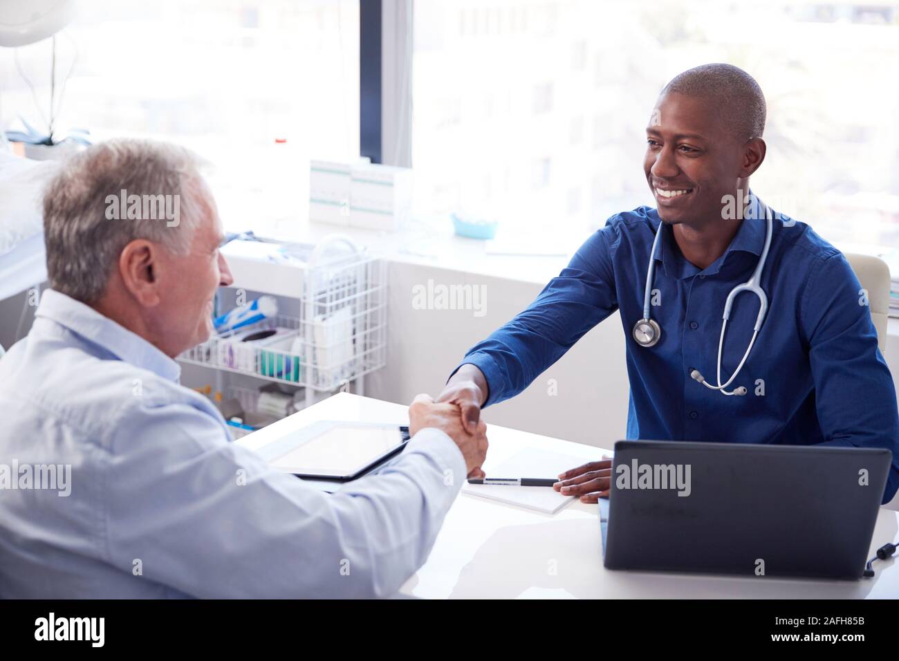 Senior Male Patient Shaking Hands With Doctor Sitting At Desk In Office Banque D'Images