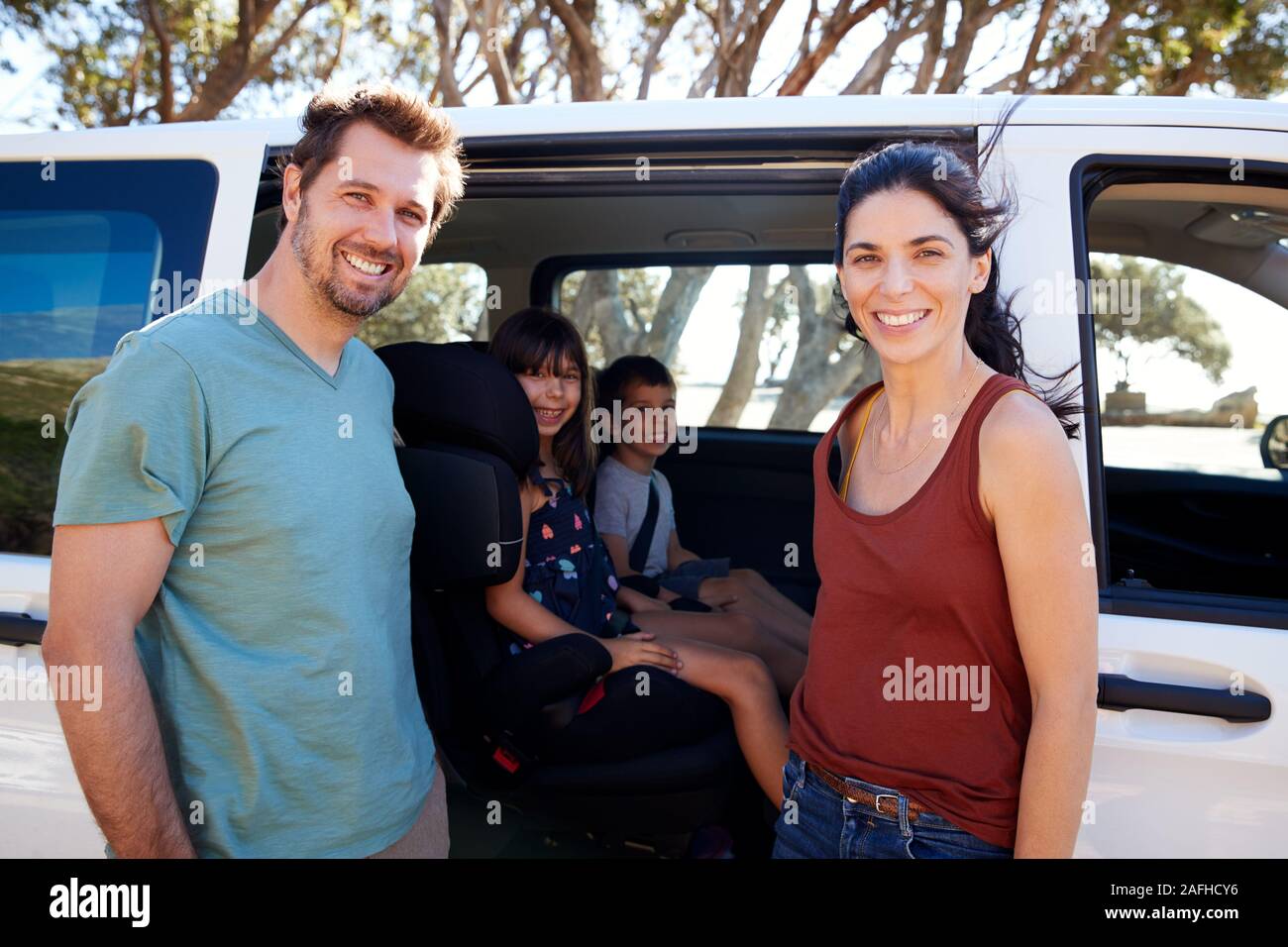 Young parents blanc debout à côté de voiture, leurs deux jeunes enfants à l'intérieur souriant pour appareil photo, Close up Banque D'Images