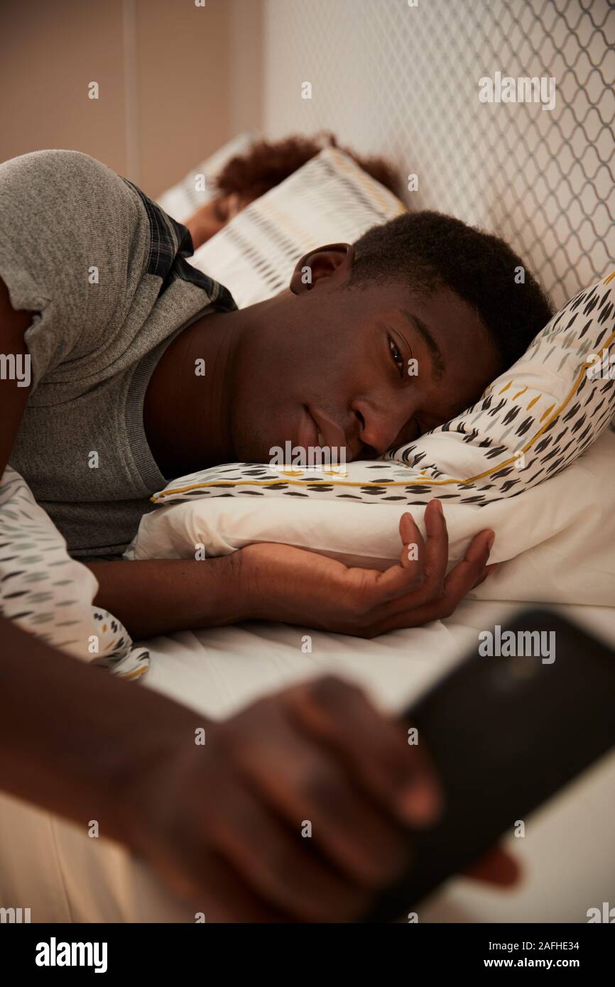 African American man millénaire à moitié endormi dans le lit, holding smartphone sa partenaire en arrière-plan, vertical Banque D'Images