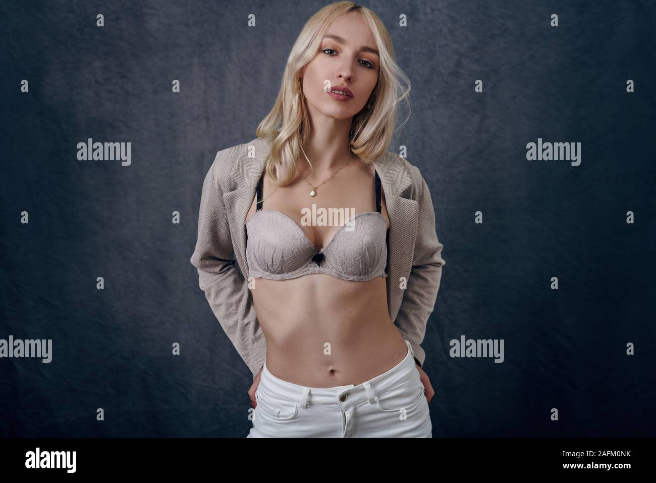 Jolie jeune femme blonde avec blazer ouvert montrant son nouveau soutien-gorge et en donnant à l'appareil photo un regard sensuel sur un arrière-plan gris studio Banque D'Images