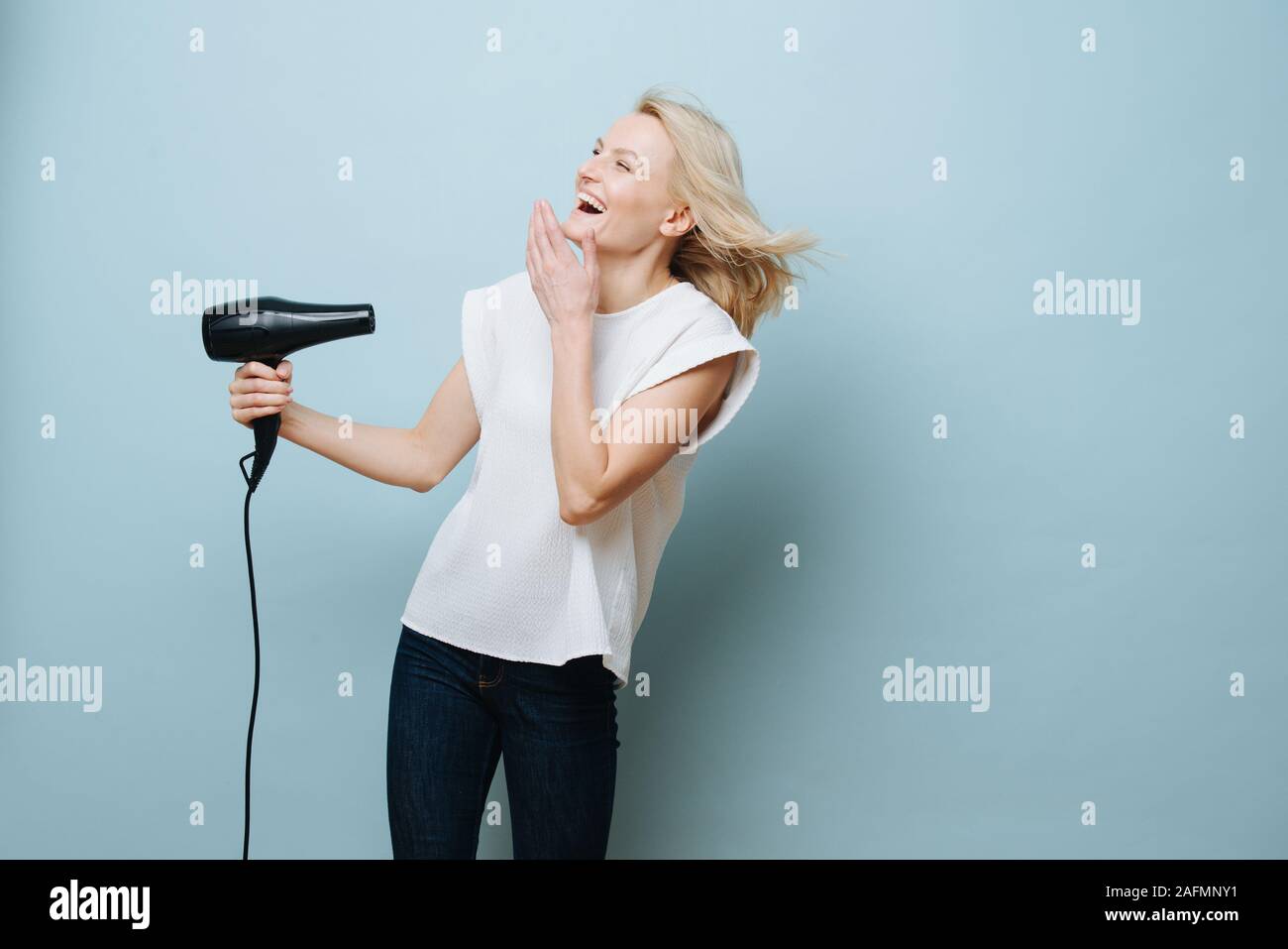 Rire blonde woman blowing elle-même dans un visage avec un sèche-cheveux plus de blue Banque D'Images