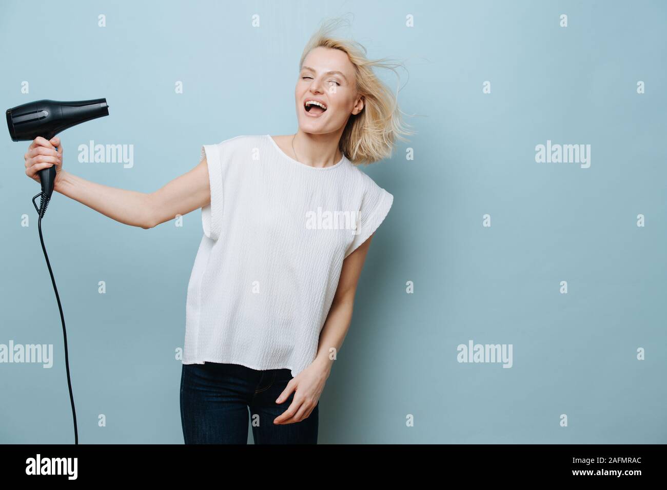 Funny blonde woman blowing elle-même dans un visage avec un sèche-cheveux plus de blue Banque D'Images