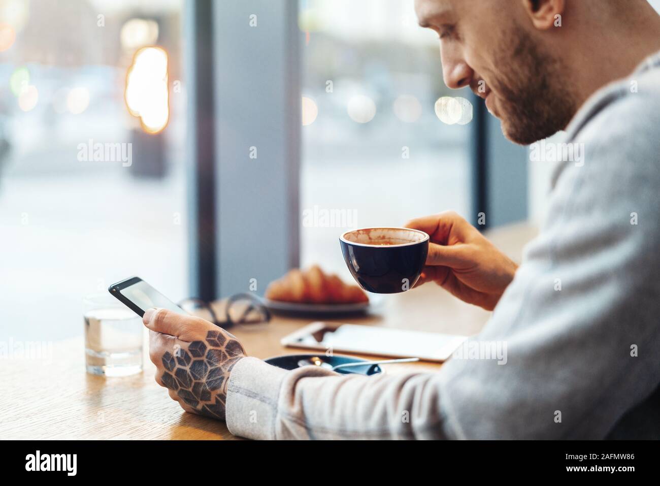Close up of Man with Tattoo à boire du café et Texting Message dans le téléphone mobile. Croissant est sur l'arrière-plan. Social Media Concept. Gadget sans fil. Co Banque D'Images