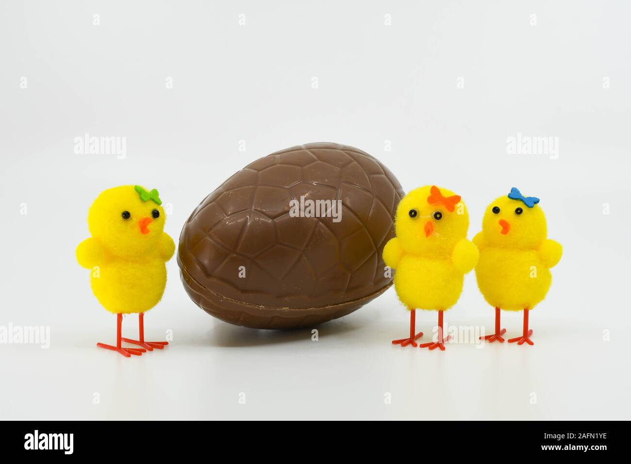 Trois poussins jaune mignon en mousse debout à côté d'un oeuf de Pâques en chocolat sur un arrière-plan uni, blanc Banque D'Images