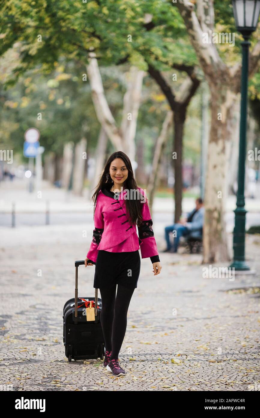 Smiling woman pulling suitcase le long trottoir bordées Banque D'Images