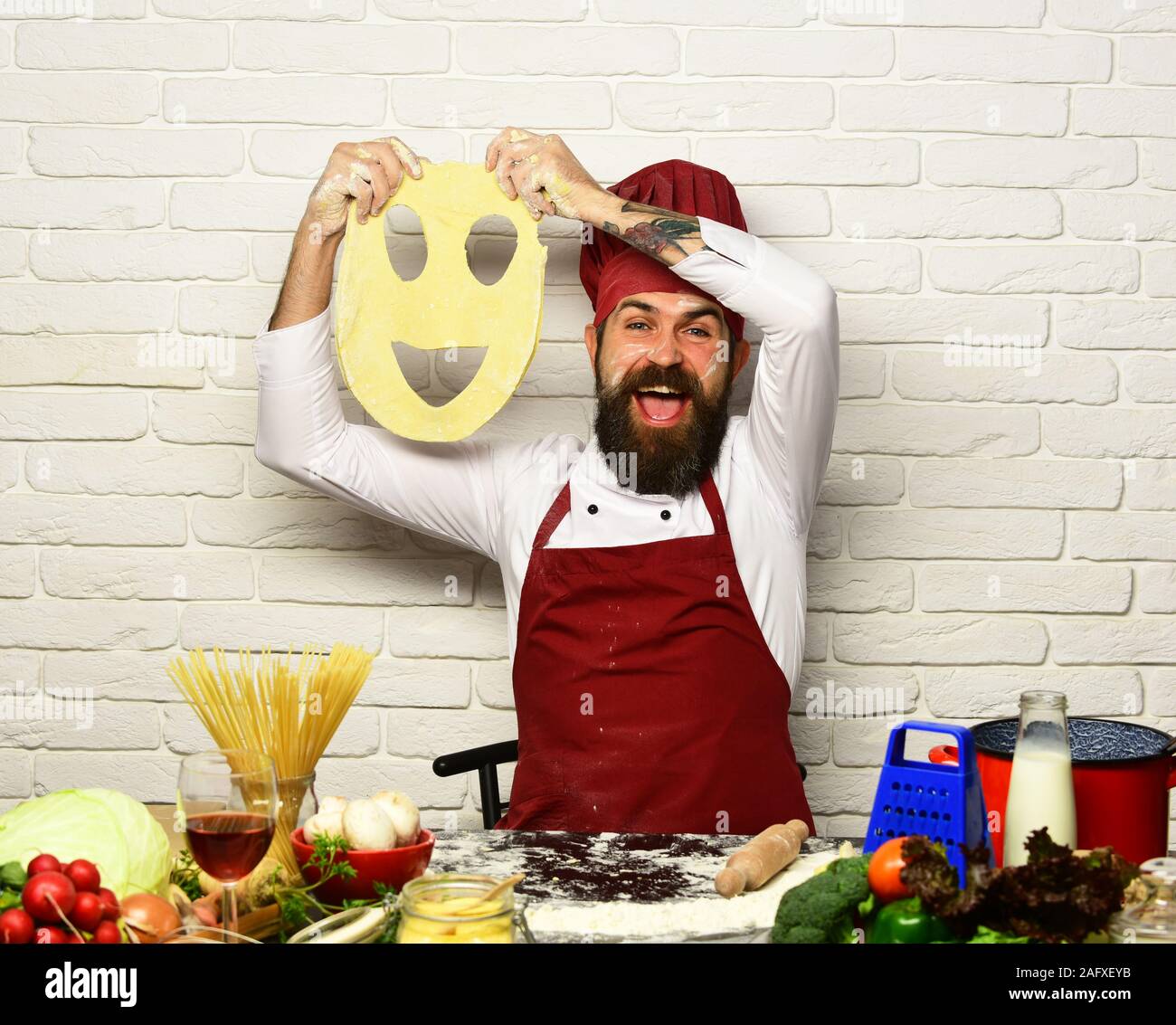 Homme à barbe est titulaire de pâte avec cut smiley sur fond de brique blanche. Cook avec visage excité en uniforme est assis par table avec des légumes et des ustensiles de cuisine. Concept de cuisine professionnelle. La pâte fait Chef Banque D'Images