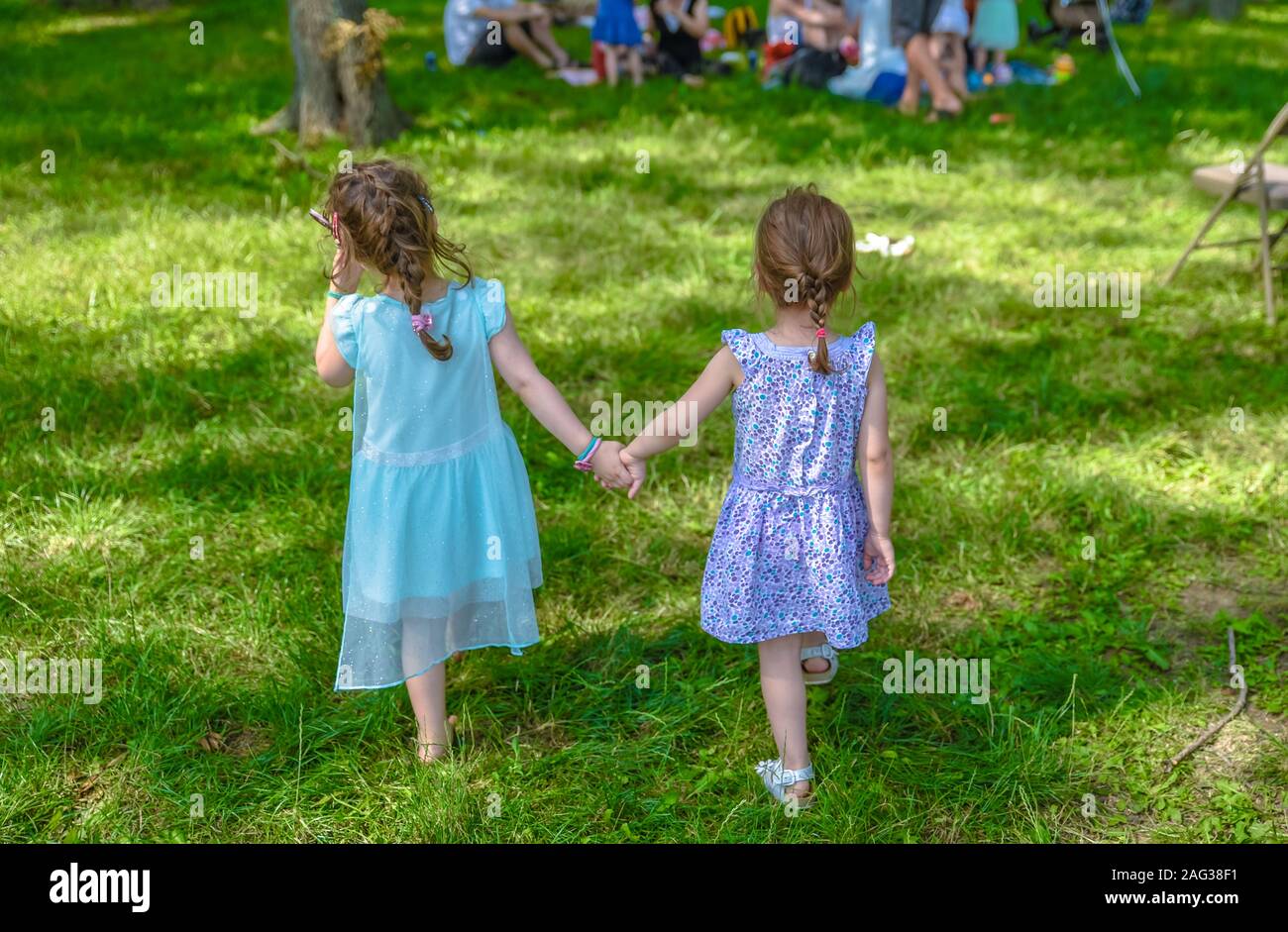 Deux jeunes filles dans des robes turquoise et violette marchant et tenant les mains dans le parc tout en approchant un groupe de personnes ayant pique-nique au parc Banque D'Images