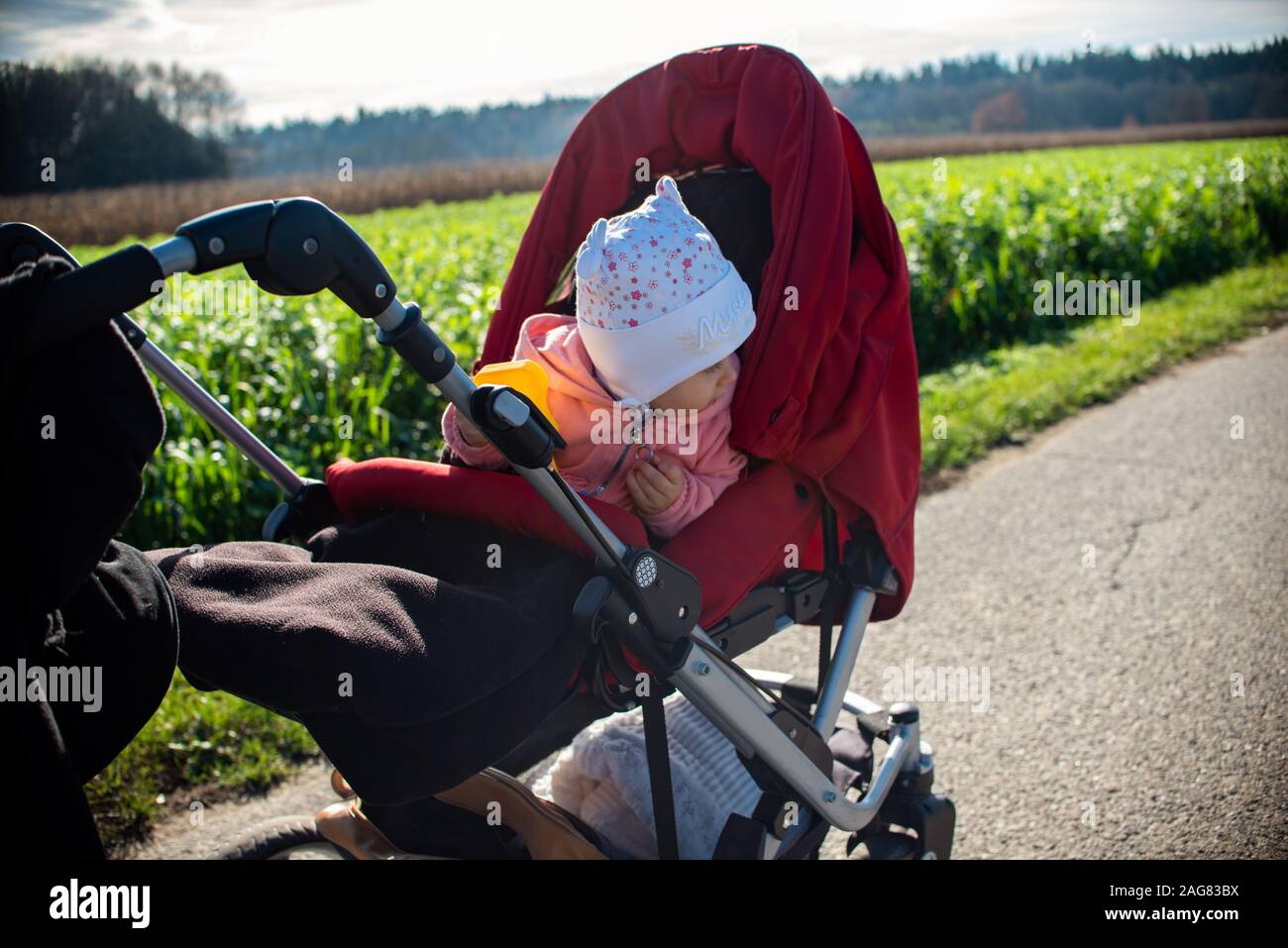 Adorable baby girl à l'extérieur en rouge poussette en vert champs sur une route, journée ensoleillée. Banque D'Images