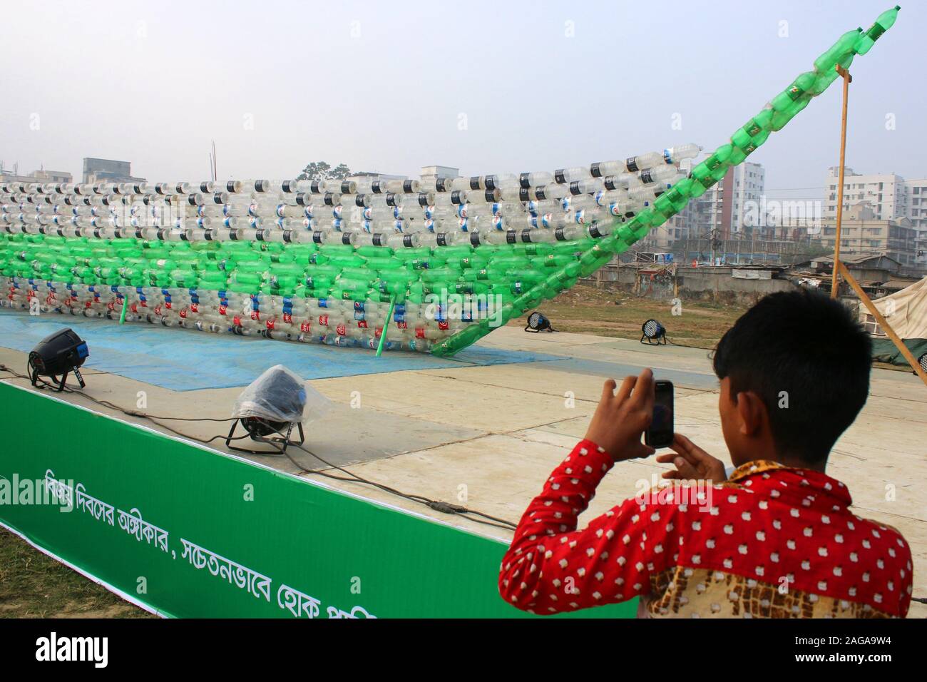 Dhaka, Bangladesh. Dec 18, 2019. Un garçon prend une photo d'un bateau fabriqué à partir de bouteilles en plastique vides lors de l'exposition.BD Nettoyer ont rassemblé 3 millions de bouteilles vides en plastique, provenant de diverses régions du Bangladesh, en hommage aux trois millions de martyrs qui ont sacrifié leur vie dans la guerre de libération de 1971. Artistes conçu divers objets, y compris une murale de São Paulo, des bateaux et la carte du Bangladesh, en utilisant les restes. Les œuvres ont été mis sur l'affichage à l'Mohakhali T&T aire de colonie à Dhaka à la veille du jour de la victoire. Credit : SOPA/Alamy Images Limited Live News Banque D'Images