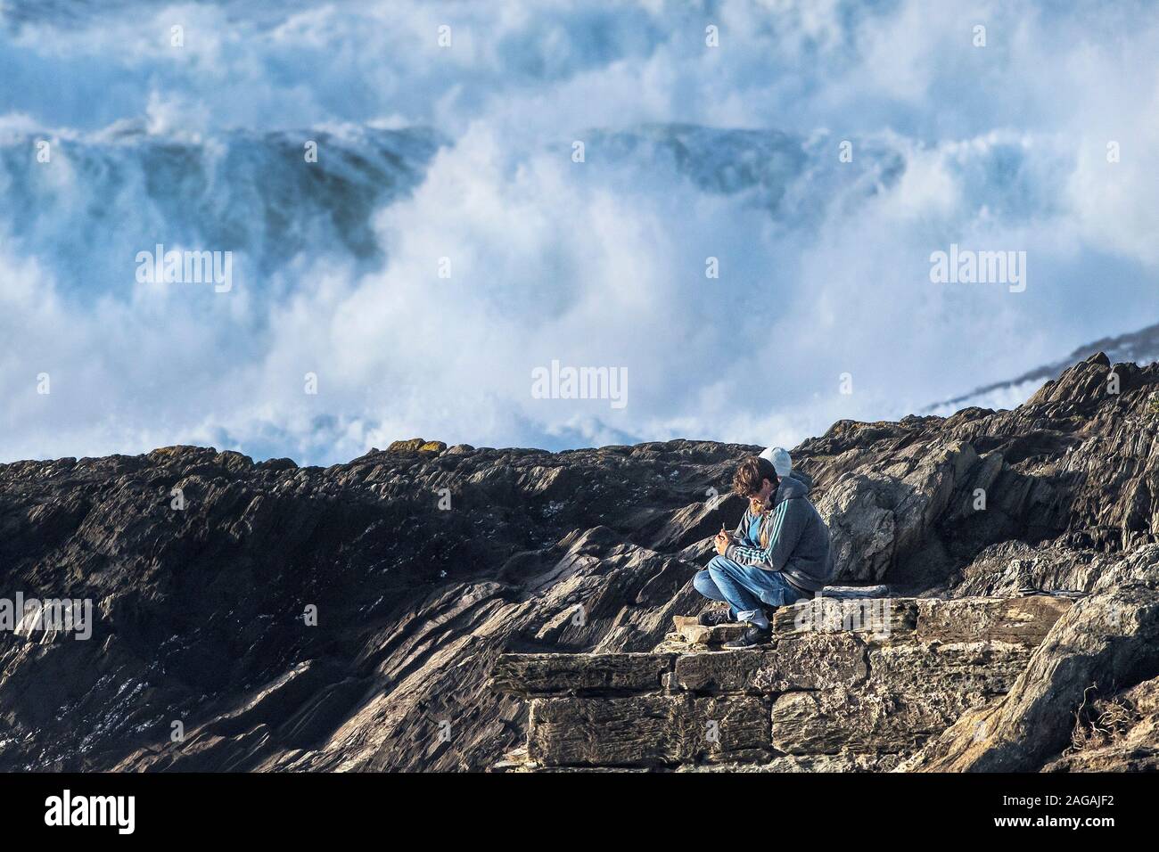 Deux jeunes gens assis dans une position dangereuse sur rochers, gros des vagues de tempête pause au fond à tête de Towan à Newquay en Cornouailles. Banque D'Images