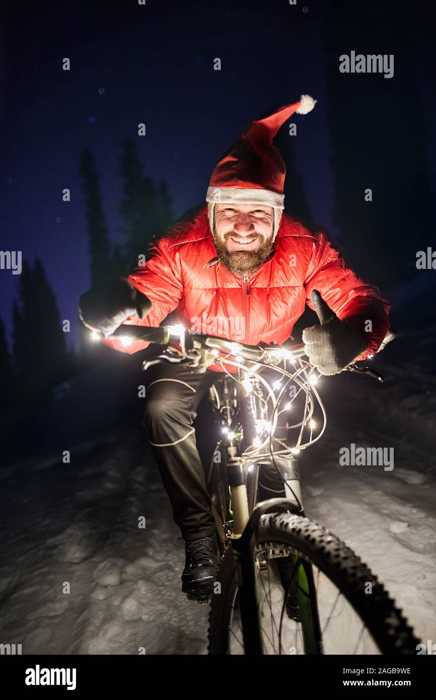 Portrait d'homme barbu en veste rouge et Noël hat le vélo à l'hiver la forêt enneigée sous ciel de nuit avec des étoiles Banque D'Images