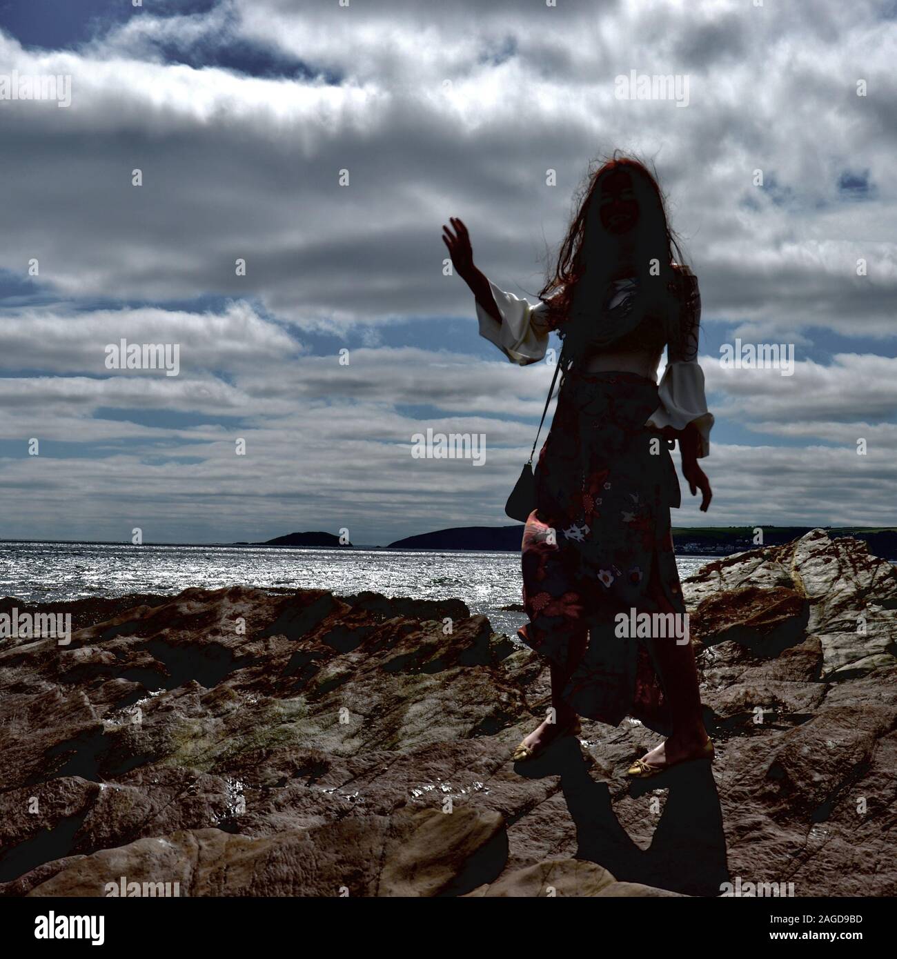 Jeune femme à la mode hippie avec de longs cheveux bruns et un pantalon évasé floral pose par la mer sur des rochers sous un cloudscape ; et rétroéclairé avec son ha Banque D'Images