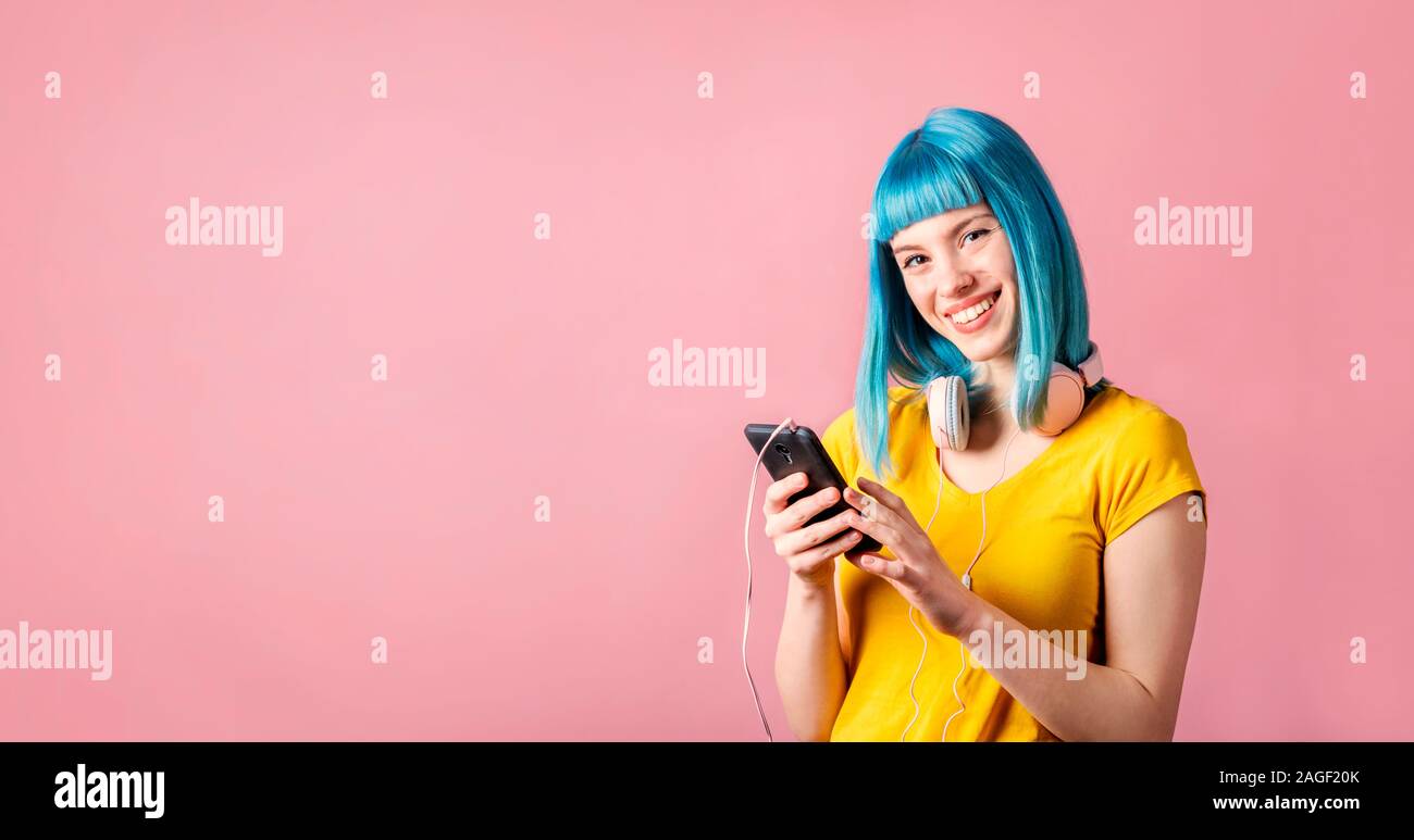 Jeune femme à la mode avec les cheveux teints en bleu holding smartphone et écouter de la musique. Hipster girl with headphones sur fond pastel neon avec copyspace Banque D'Images