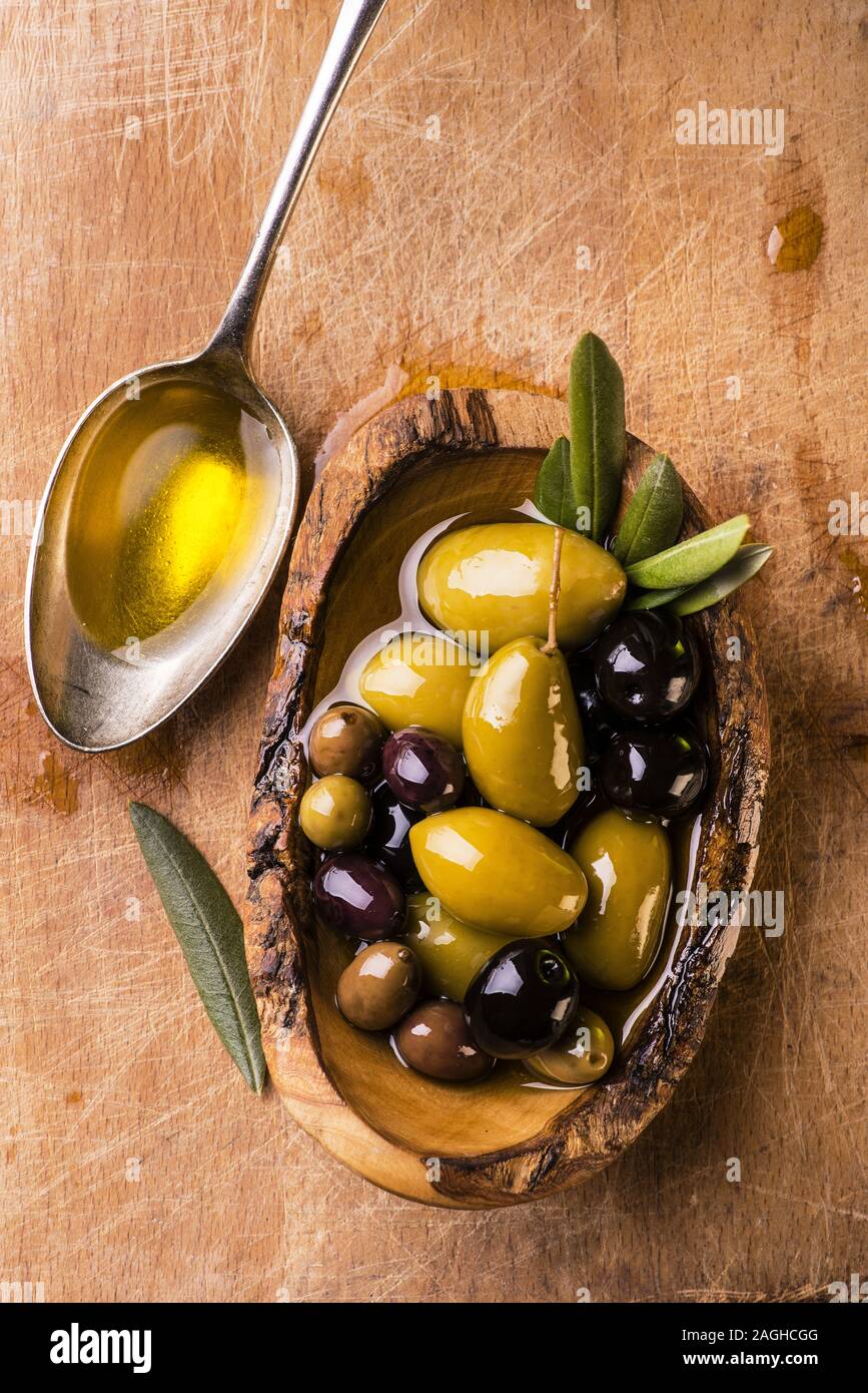 Sur la table de bois rustique, un bol avec différents types d'olives et une cuillère pleine d'huile Banque D'Images