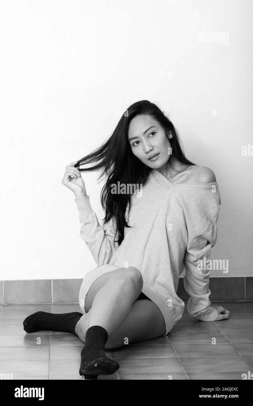 Studio shot of young beautiful Asian woman assis sur le plancher tout en jouant avec ses cheveux et la pensée Banque D'Images Studio shot of young beautiful Asian woman assis sur le plancher tout en jouant avec ses cheveux et la pensée Banque D'Images
