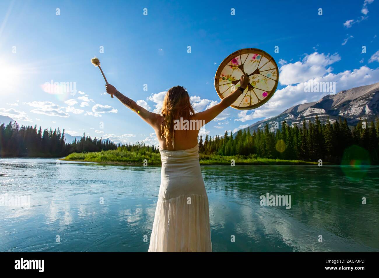La culture autochtone traditionnelle répond à mère nature comme un compte femme portant une robe d'été blanche détient tambour sacré et du batteur par une rivière Banque D'Images