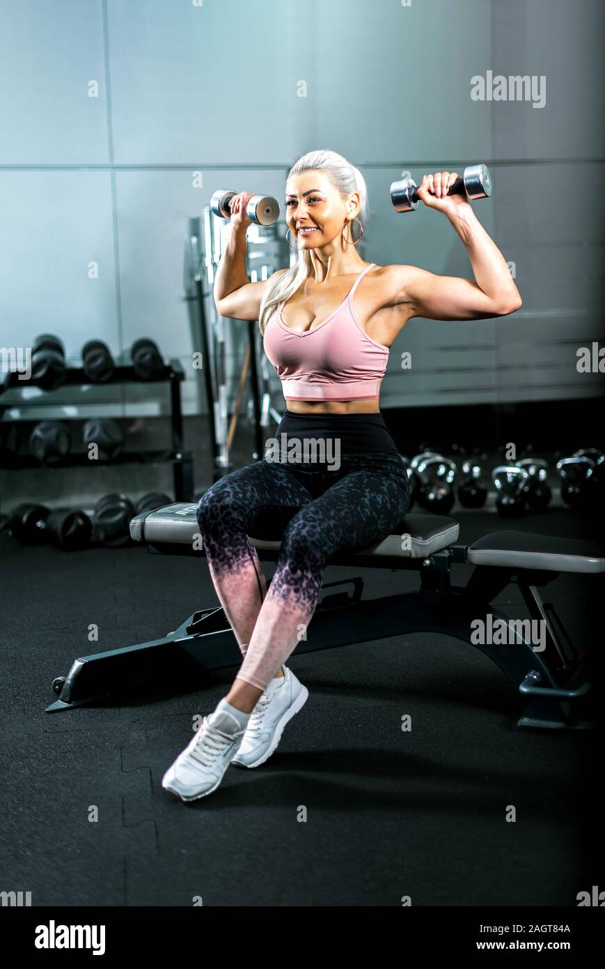 Blonde woman doing biceps haltères assis sur un banc dans la salle de remise en forme moderne. La tonalité de l'image. Banque D'Images