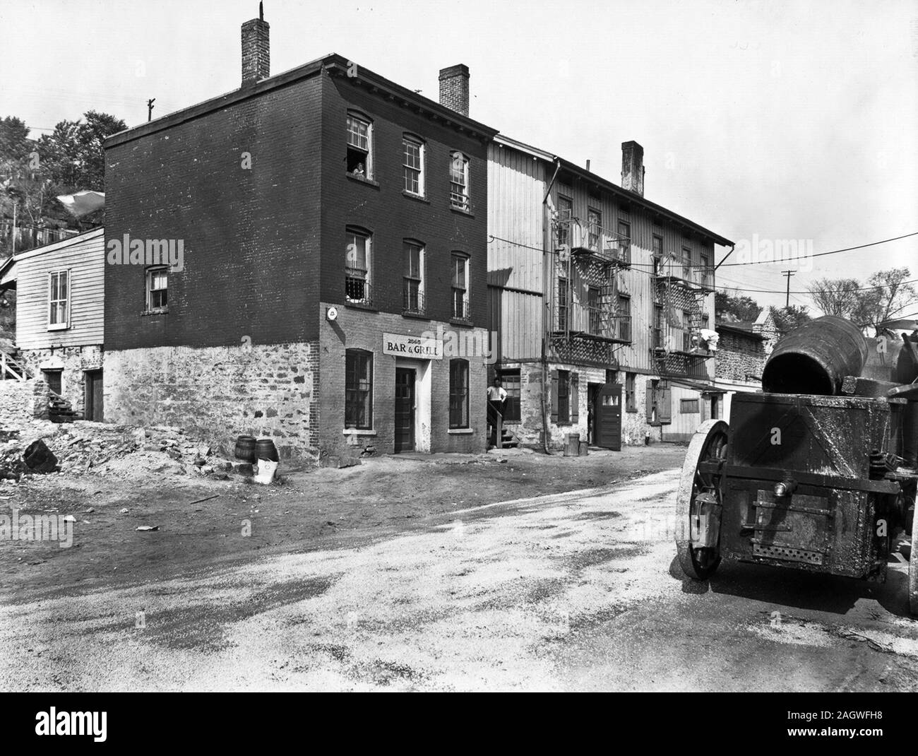 Bar et grill et autres édifices construits dans la rue le long de la colline, l'homme a l'air sur des étapes, fille de fenêtre au-dessus, véhicule avec barils, droite. Banque D'Images
