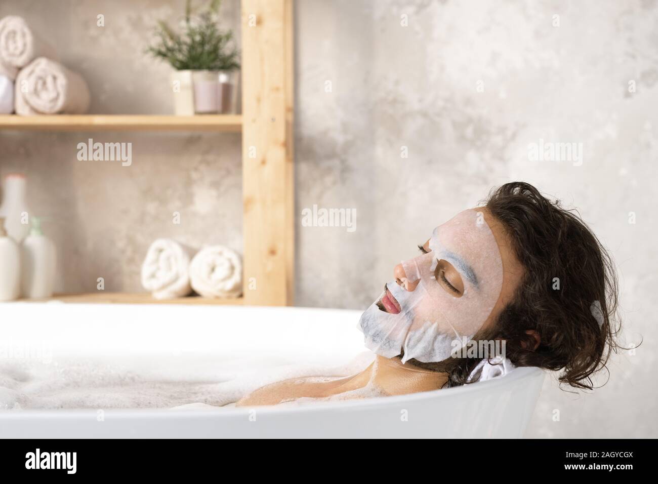 Jeune homme serein et détendu avec masque de visage appréciant bain chaud avec de la mousse Banque D'Images