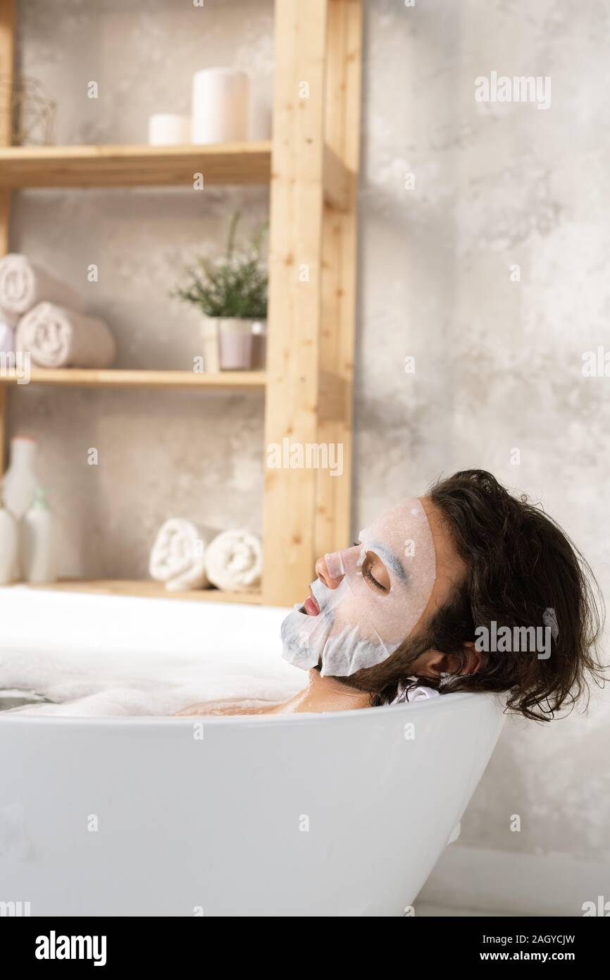 Ambiance jeune homme ayant masque de visage tout en se trouvant dans la baignoire avec de l'eau chaude et la mousse Banque D'Images