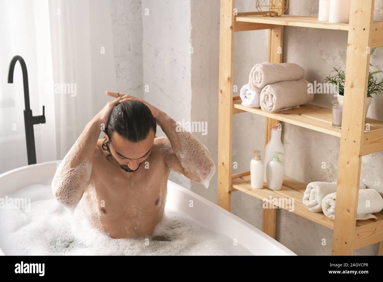 Jeune homme musclé humide laver ses cheveux tout en ayant baignoire avec mousse Banque D'Images