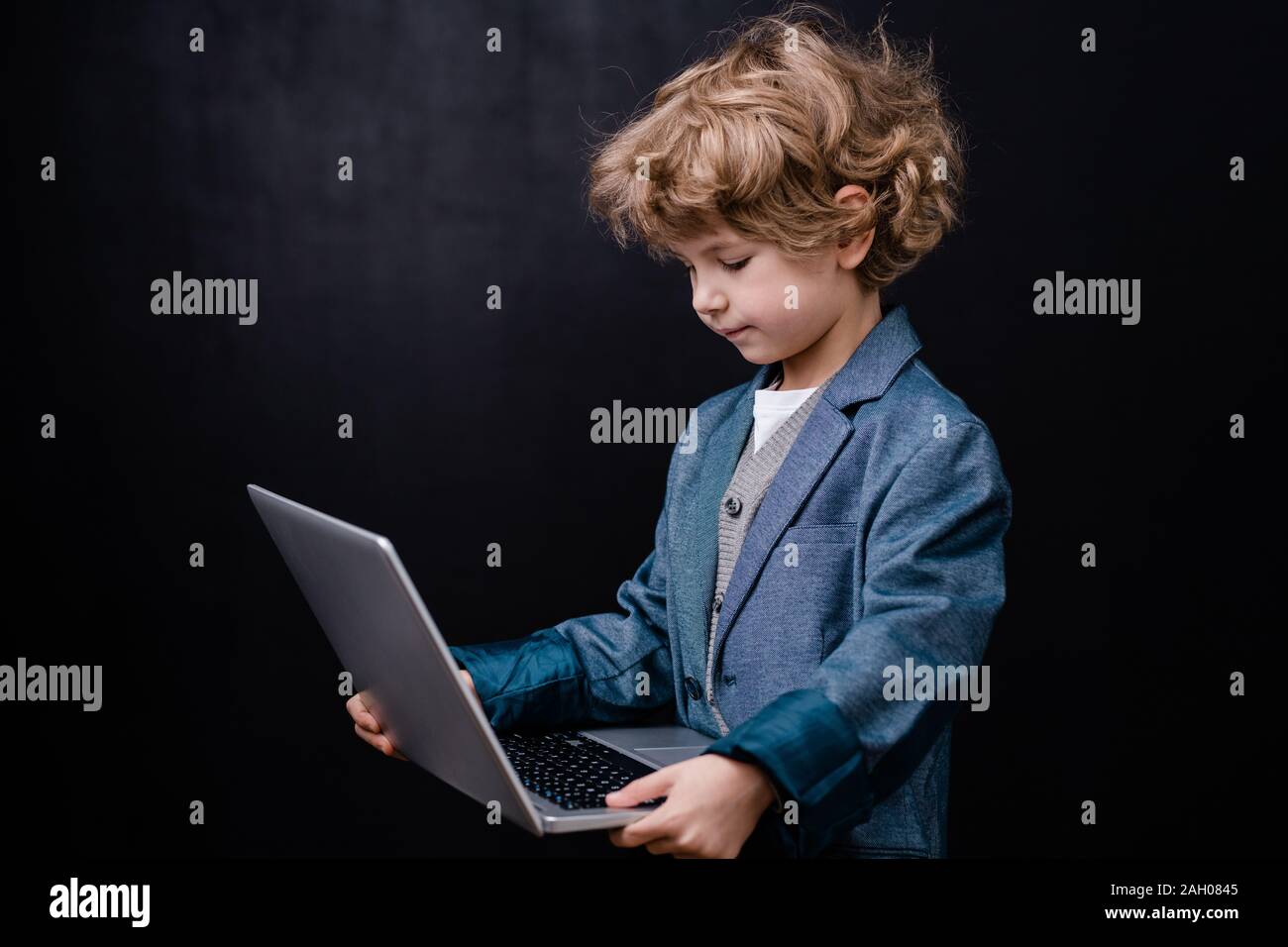 Clever elementary schoolboy holding laptop en face de lui-même en surfant sur le net sur fond noir Banque D'Images
