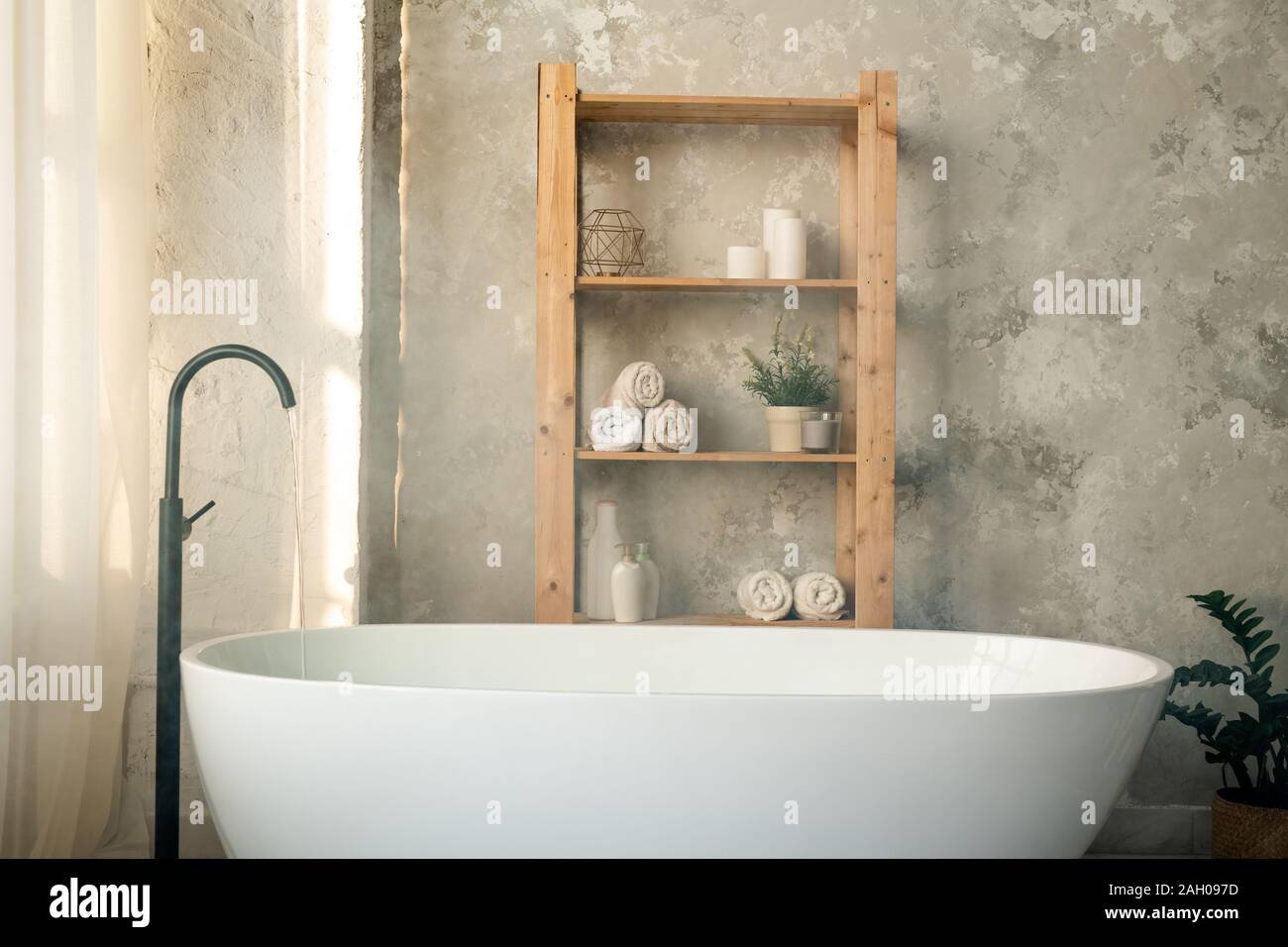 Grande baignoire blanc porcelaine et étagères en bois avec des serviettes roulées, pots en plastique et des bougies contre mur gris dans la salle de bains Banque D'Images