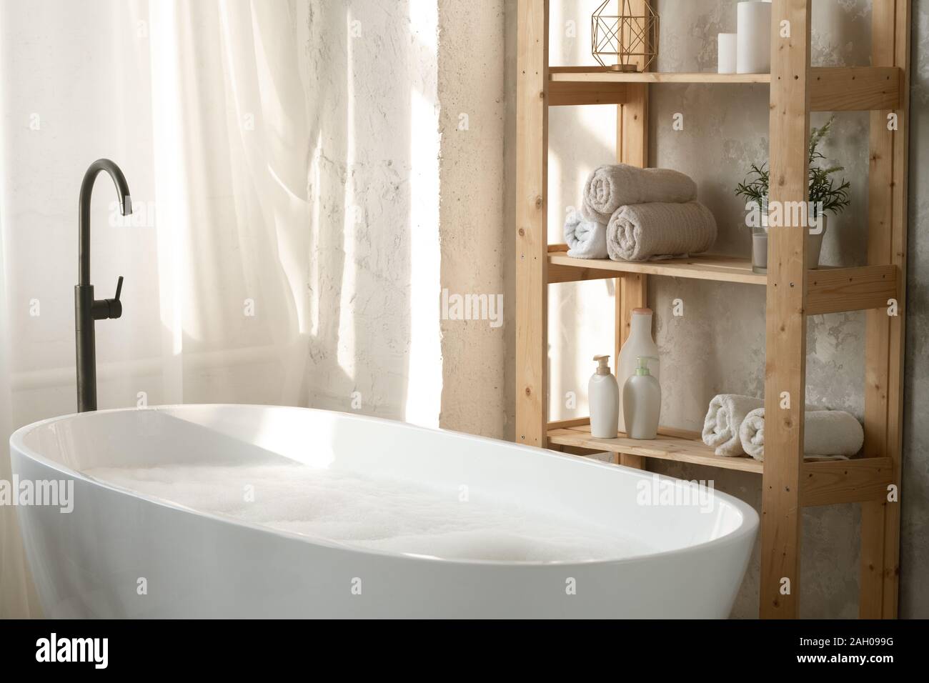 Grand blanc porcelaine baignoire remplie d'eau et de mousse par des étagères en bois avec des serviettes roulées et pots en plastique contre le mur dans la salle de bains Banque D'Images