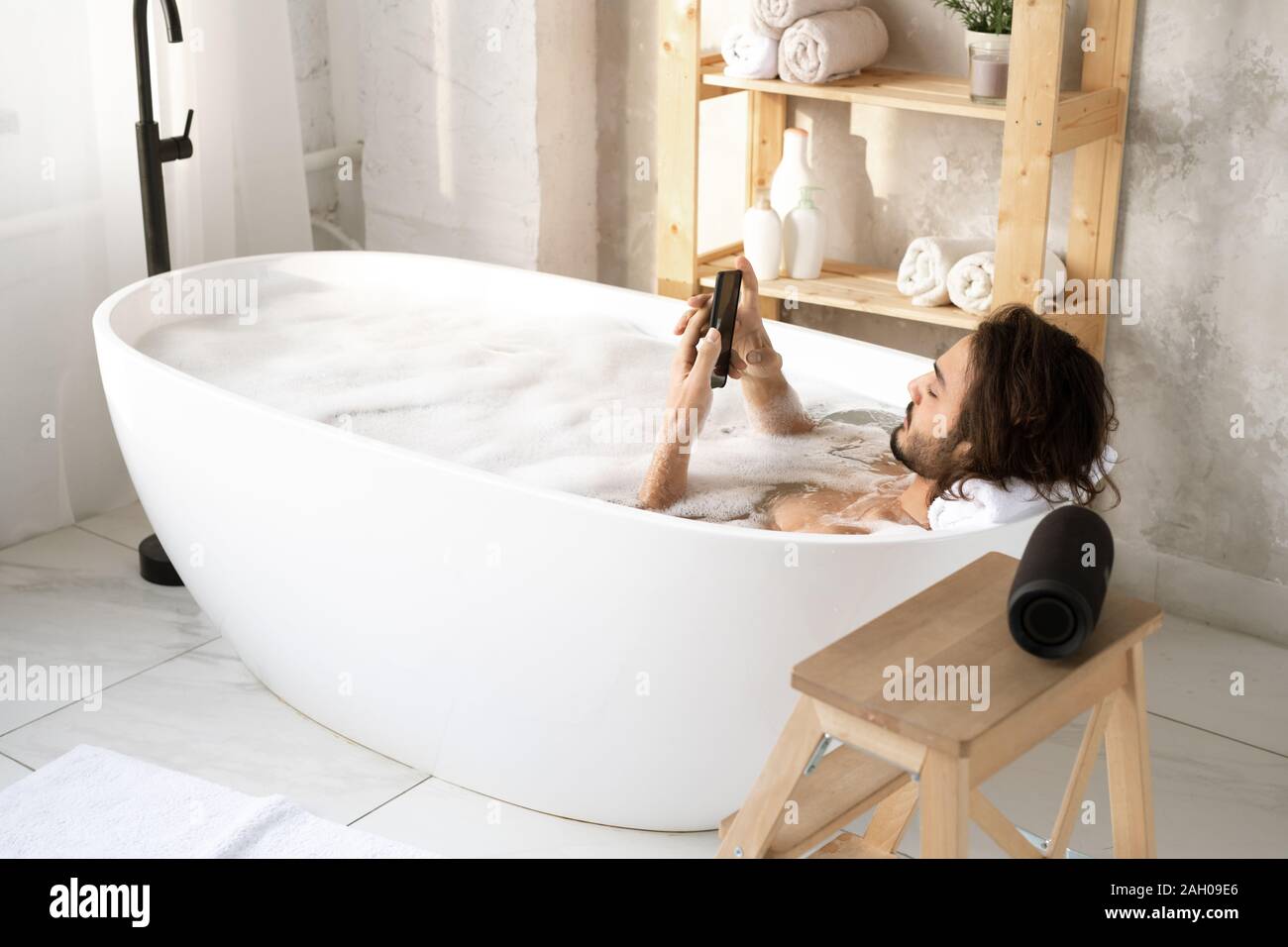 Young man using smartphone reposant tout en étant allongé dans la baignoire remplie d'eau et de mousse dans le coin d'une salle de bains privative Banque D'Images