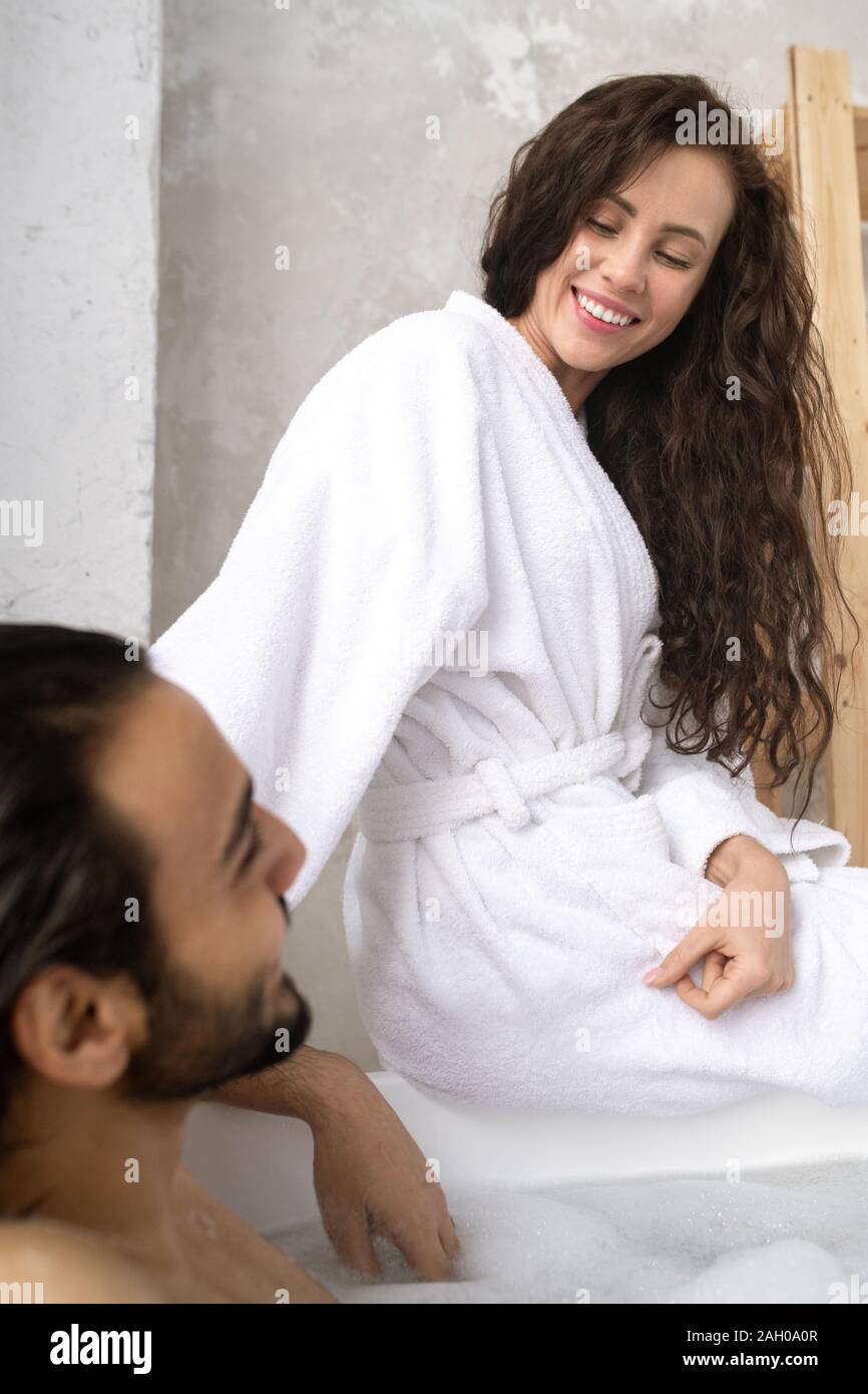 Happy young smiling woman in white peignoir de parler à son mari ayant baignoire Banque D'Images