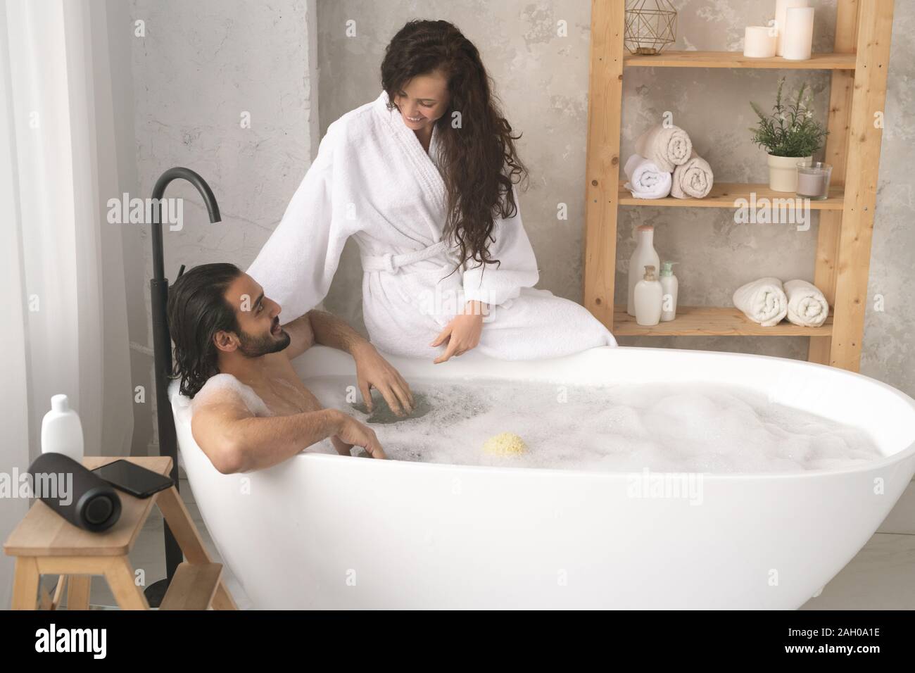 Happy young woman in bathrobe de parler à son mari enjoying bath avec mousse Banque D'Images