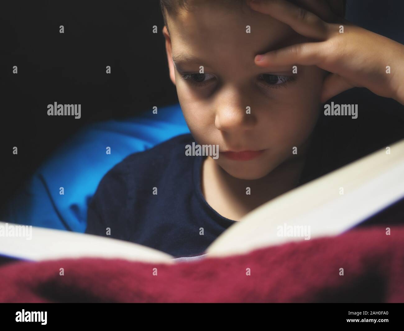 Petit enfant, 7 ans garçon lisant un livre à l'intérieur à dim spot light Banque D'Images